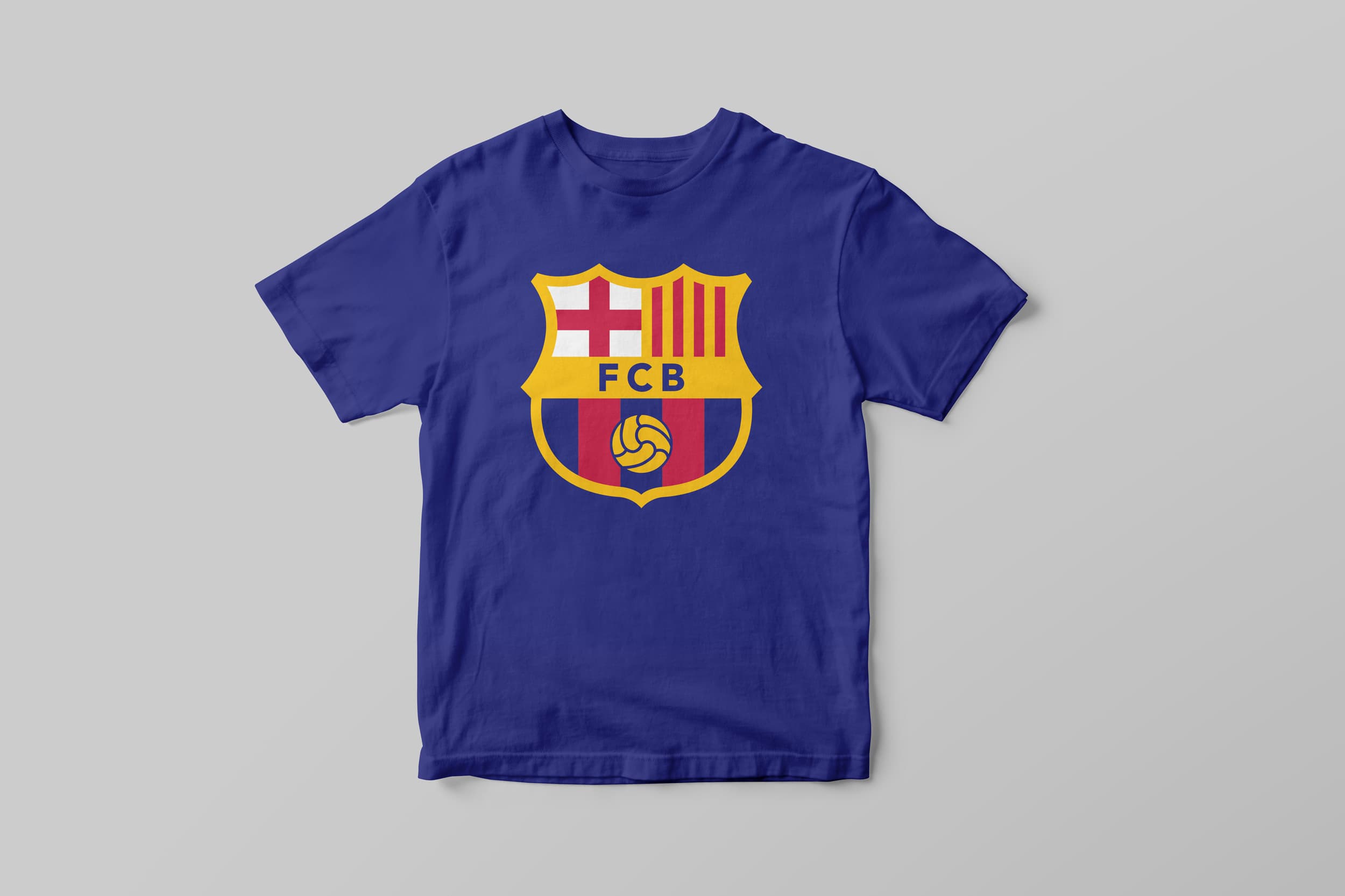 A royal blue T-shirt features a prominent shield-shaped logo at the center. The logo has "FCB" in the middle, a red cross on the upper left, red and yellow stripes on the upper right, and a soccer ball at the bottom. The background is light gray.