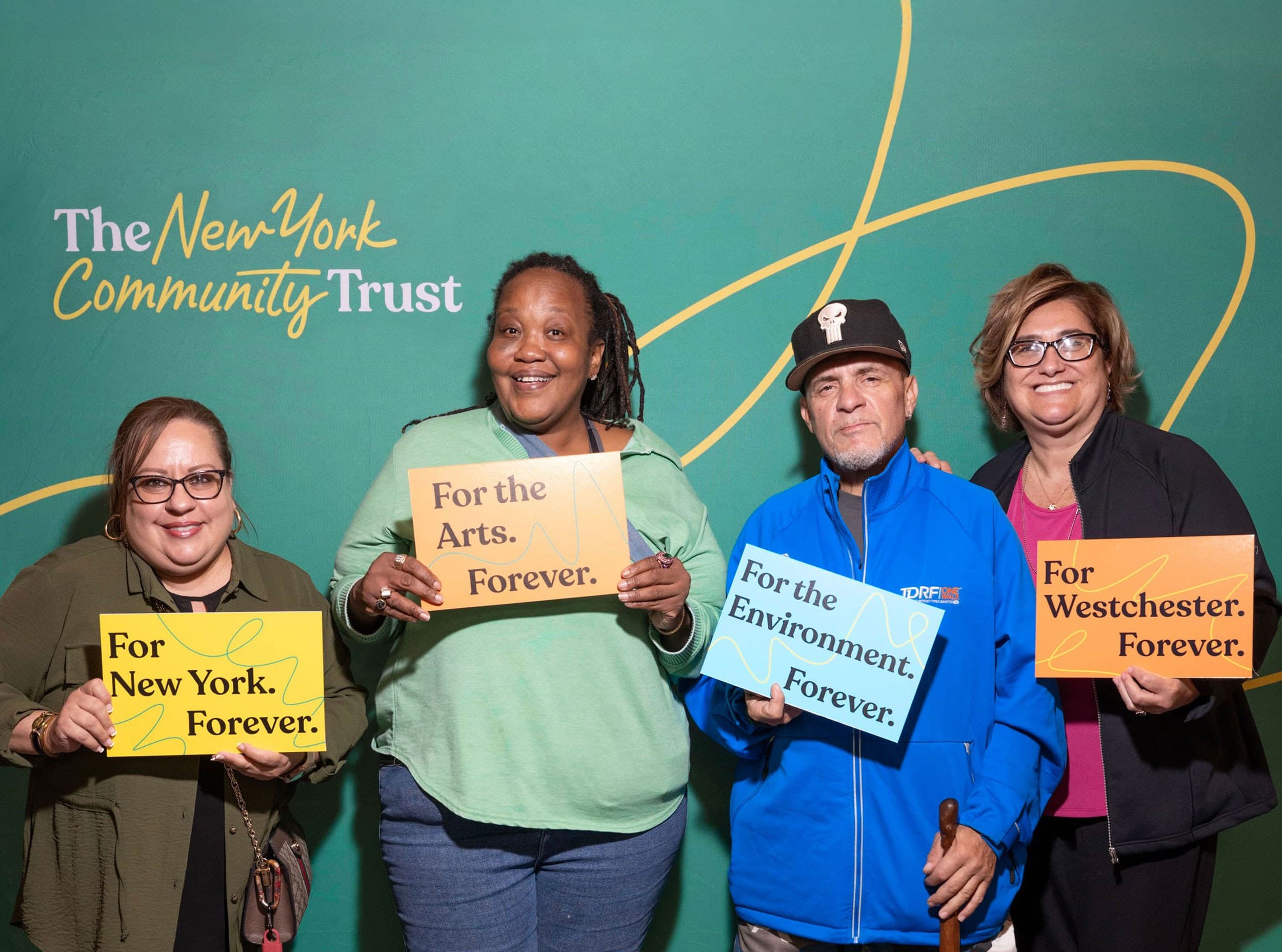 Four people stand smiling in front of a green New York Community Trust backdrop, each holding signs reading: "For New York. Forever.", "For the Arts. Forever.", "For the Environment. Forever.", and "For Westchester. Forever.