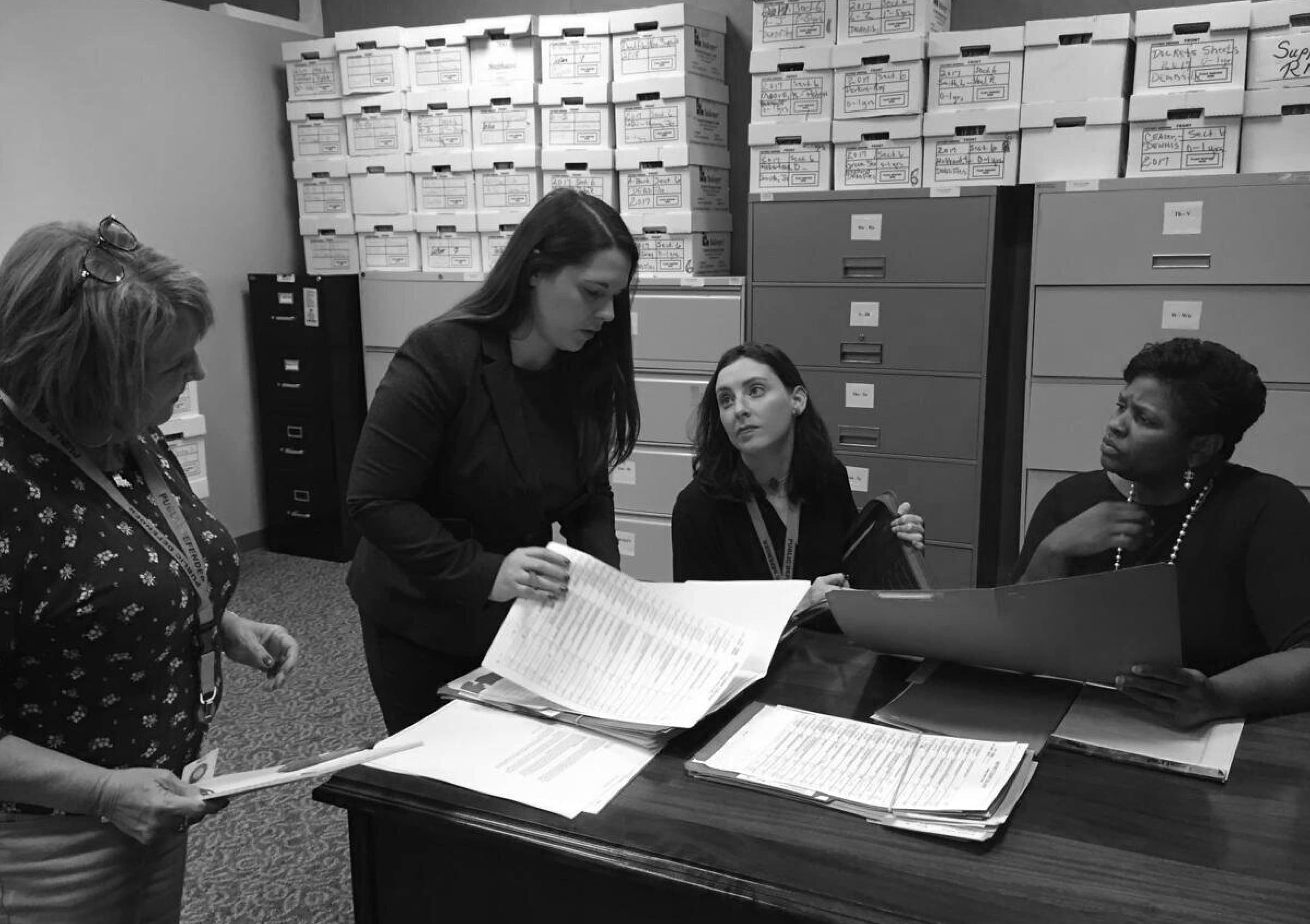 Four women are gathered around a table in an office. Three are sitting, and one is standing and holding documents. The background has shelves filled with labeled boxes and filing cabinets. They appear to be engaged in a discussion or working on paperwork.