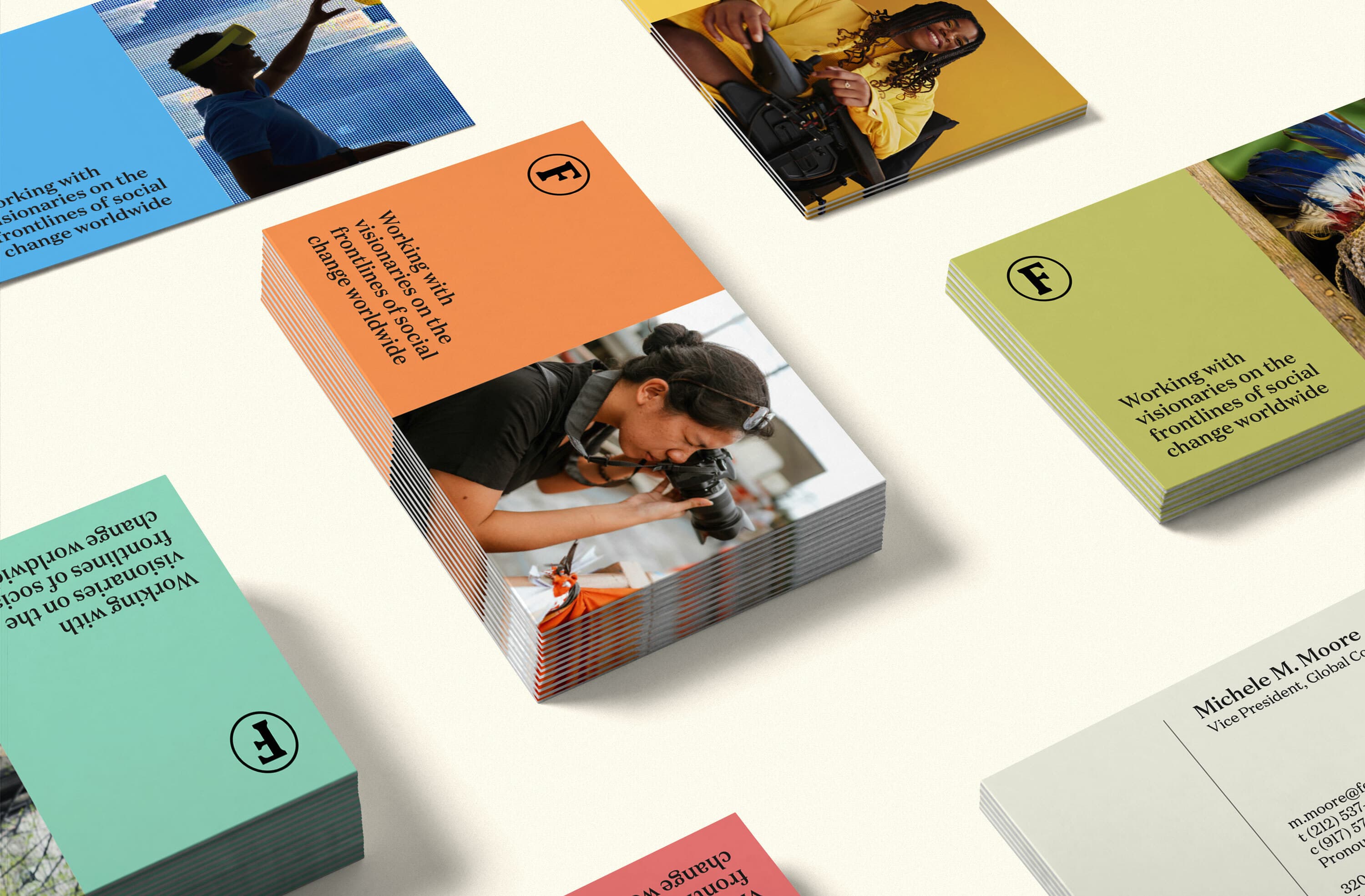 Stacks of color-coded brochures on a light surface, some depicting images of people engaged in various activities like photography and holding gears. Text on the brochures reads, "Working with frontline communities to change the world" and features a logo with an "F.