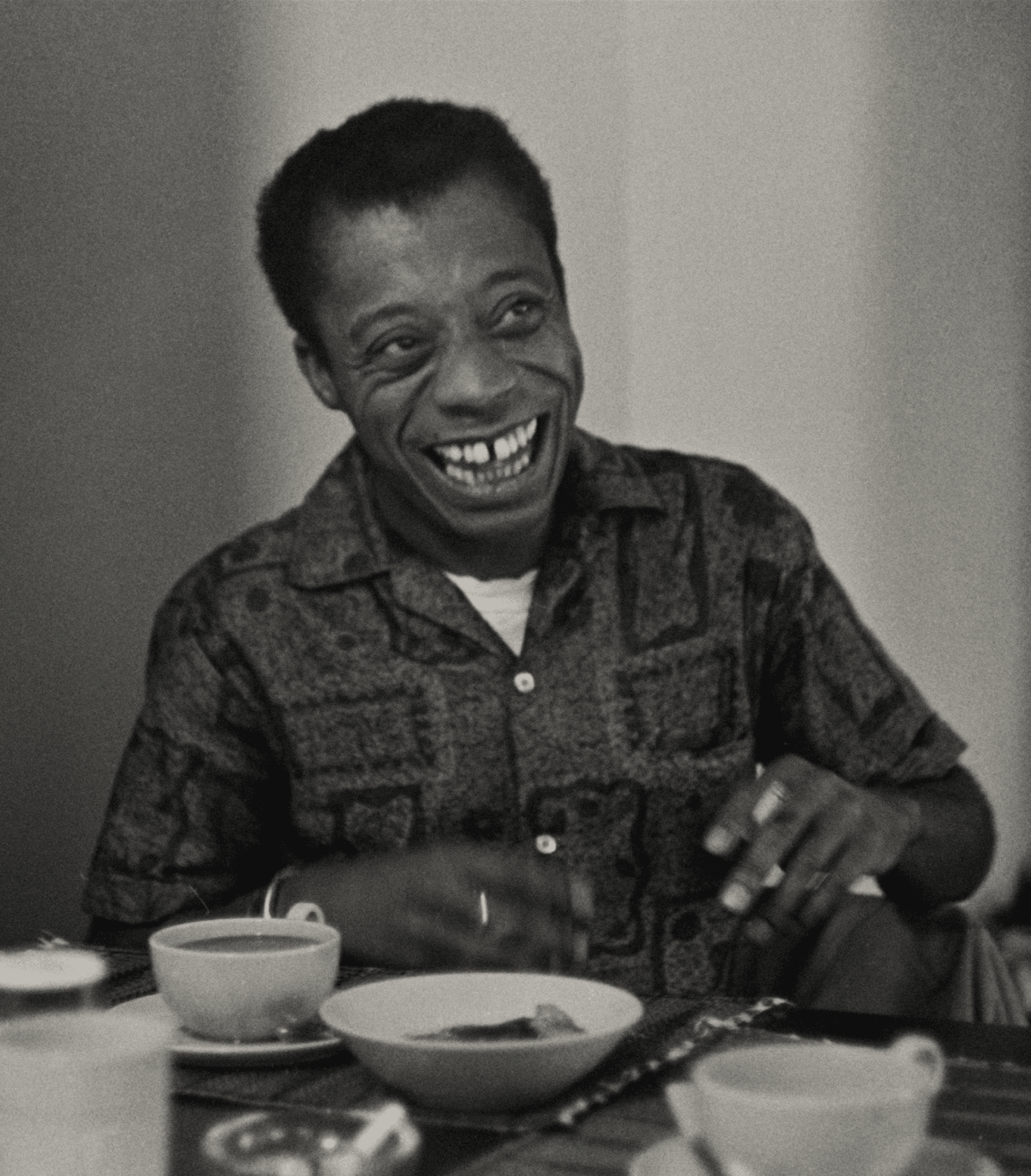 A person with a wide smile sits at a table with several cups and a bowl. They are wearing a patterned shirt and have short, dark hair. The setting appears to be indoors, possibly a café or dining area. The person looks happy and engaged in a pleasant moment.