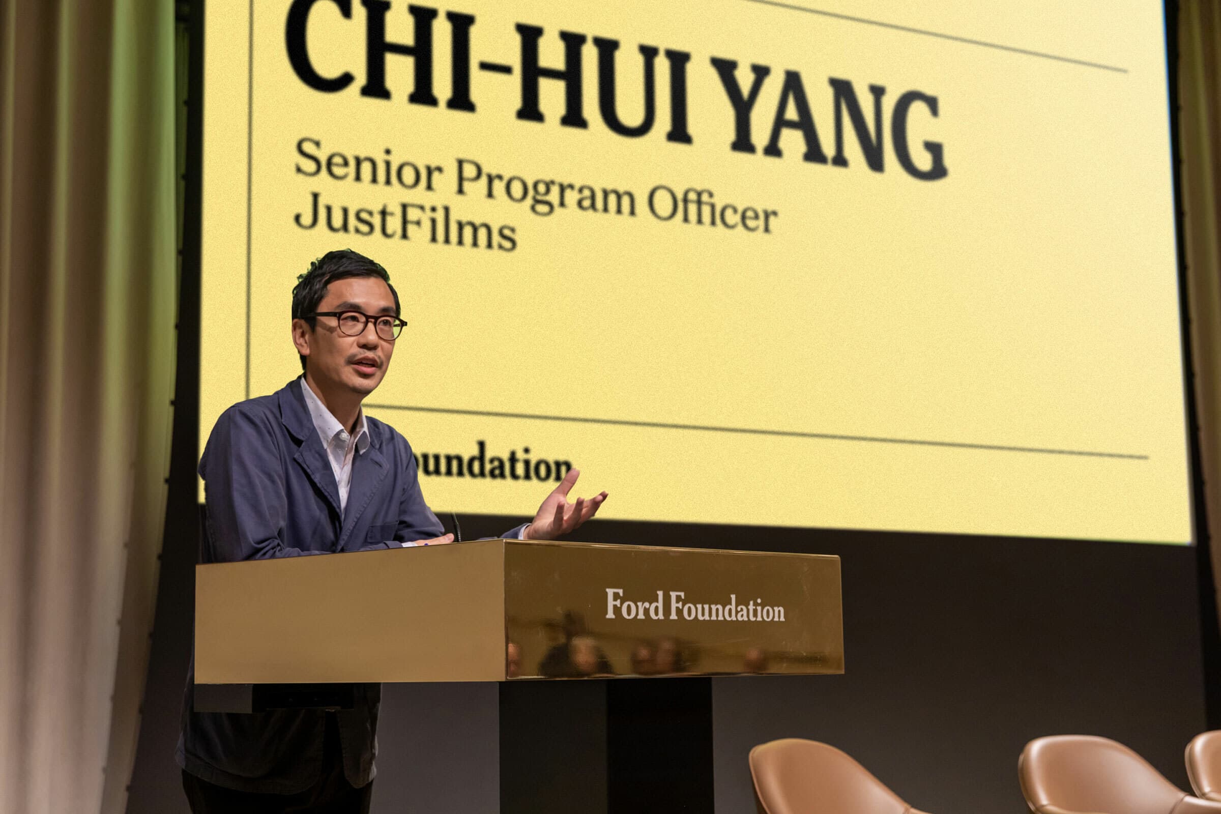 Chi-Hui Yang, identified as a Senior Program Officer for JustFilms, stands at a podium with "Ford Foundation" labeled on it. He is speaking with a large screen behind him displaying his name and title in large text.