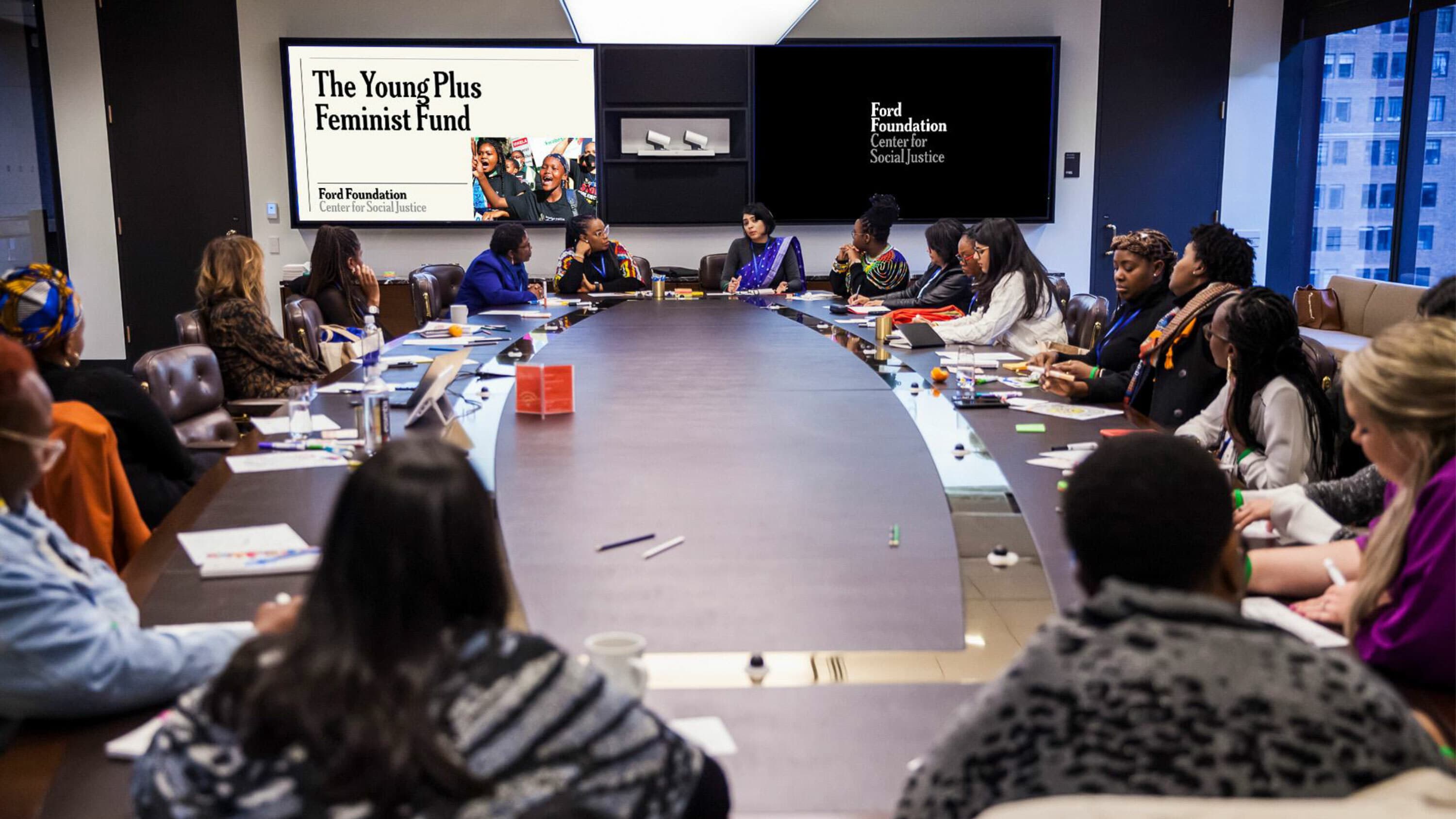 A diverse group of people are seated around a large conference table in a modern meeting room. Two large screens display "The Young Plus Feminist Fund" and "Ford Foundation Center for Social Justice." Pens, papers, and laptops are scattered on the table.