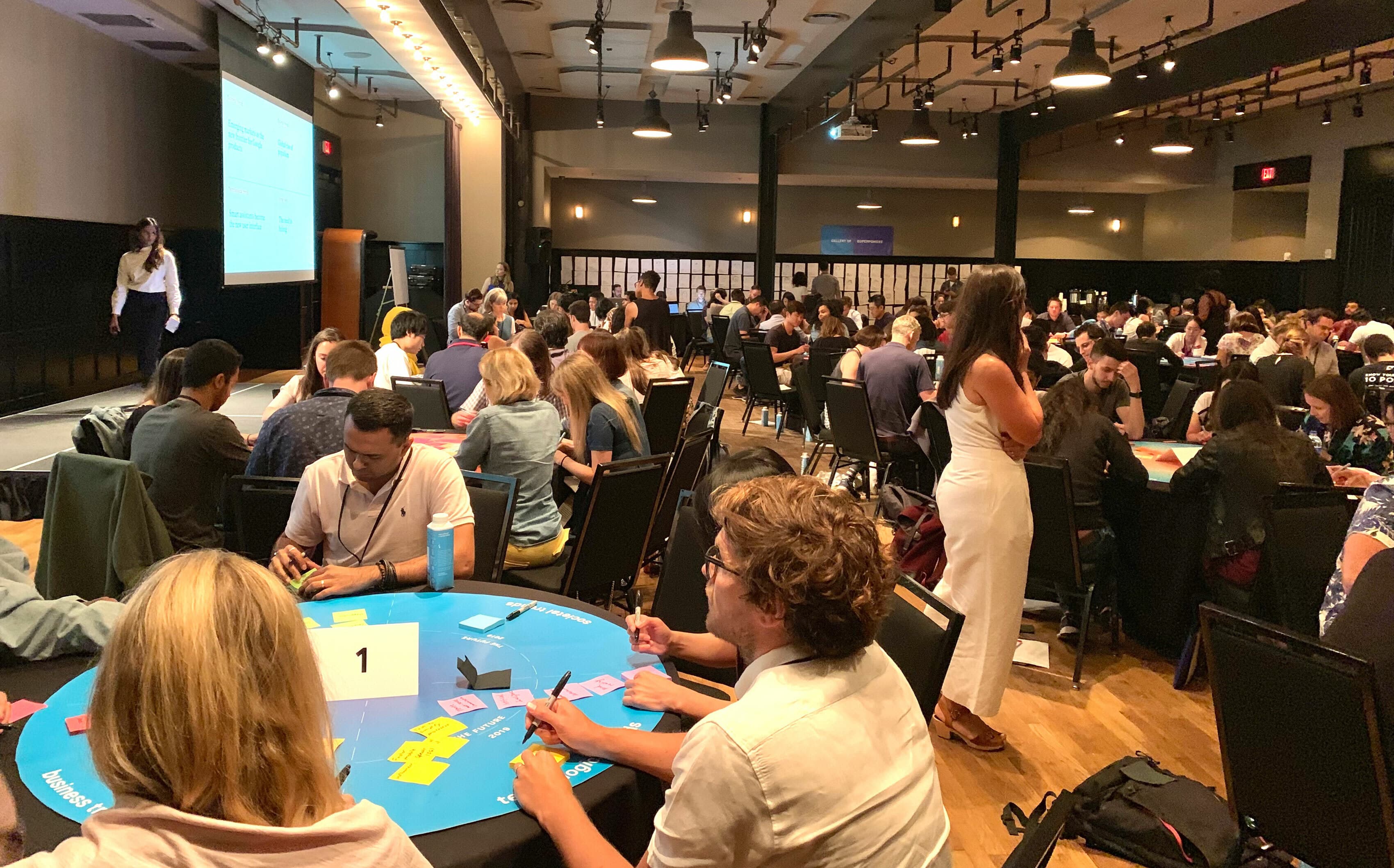 A large group of people gathered in a spacious conference room with tables arranged in rows. Attendees are engaged in discussions and activities, with some writing on colorful sticky notes. A presenter stands near a screen displaying a presentation.