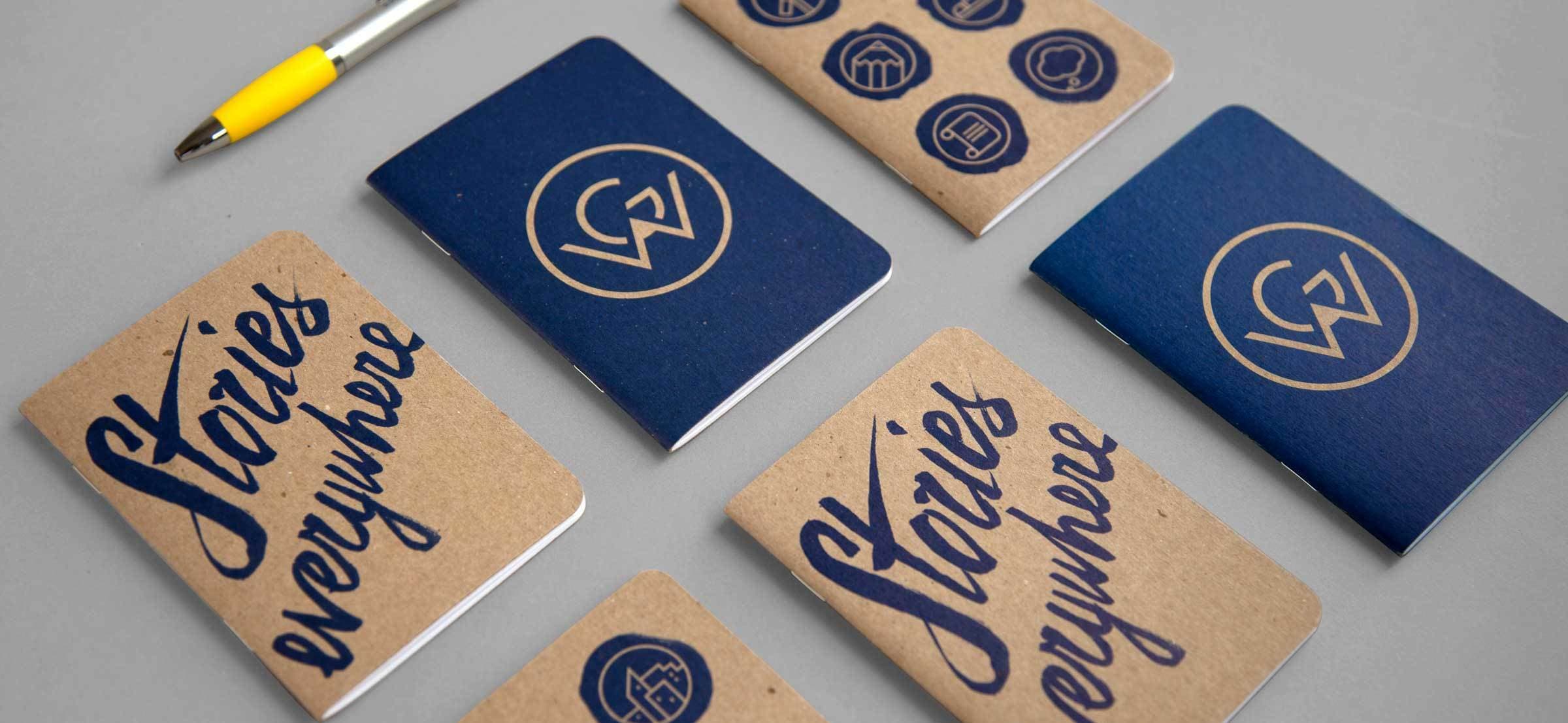 A set of notebooks is laid out on a gray surface. Some have brown covers with the words "Stories everywhere" in blue script, while others have blue covers with various icons or circular logos. A yellow pen is positioned at the top left of the image.