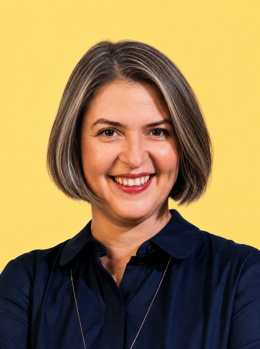 Smiling woman with short brown and gray hair, wearing a dark blue collared shirt, standing in front of a plain yellow background.