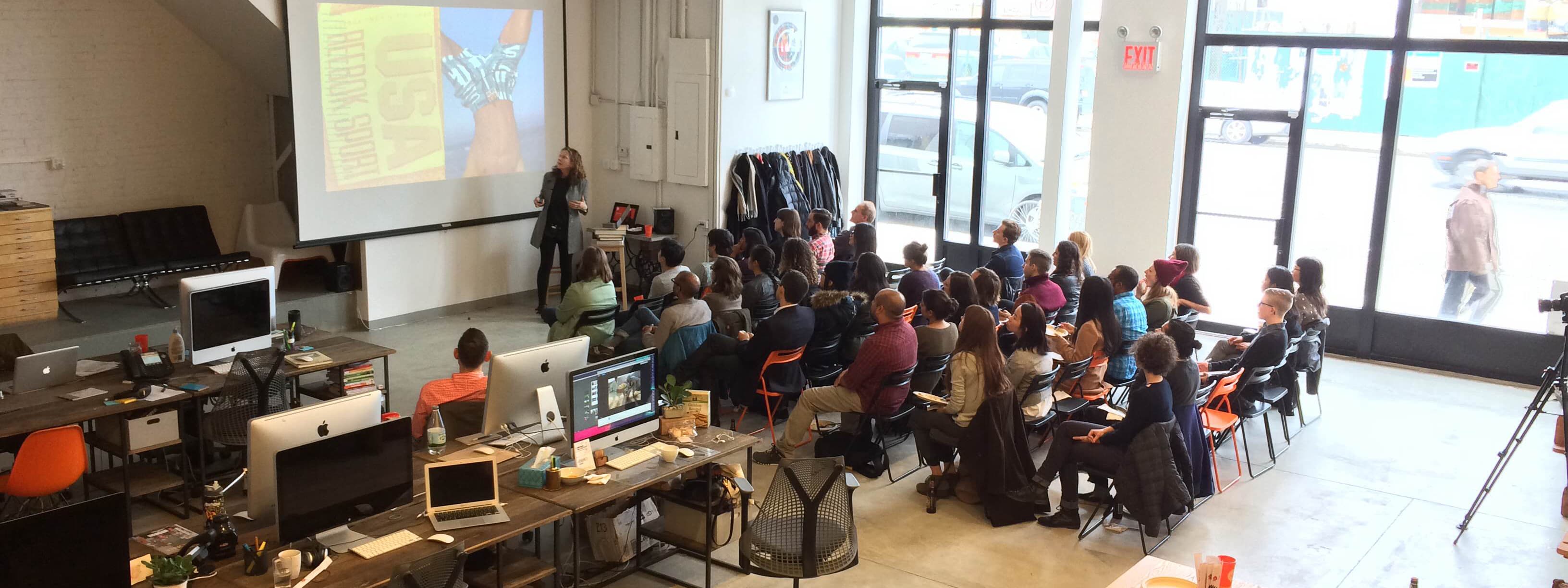 A group of people seated in a modern, open office space, attentively watching a presentation projected on a screen. The presenter stands near the screen with a microphone. Computers and office supplies are visible on desks, and large windows let in natural light.