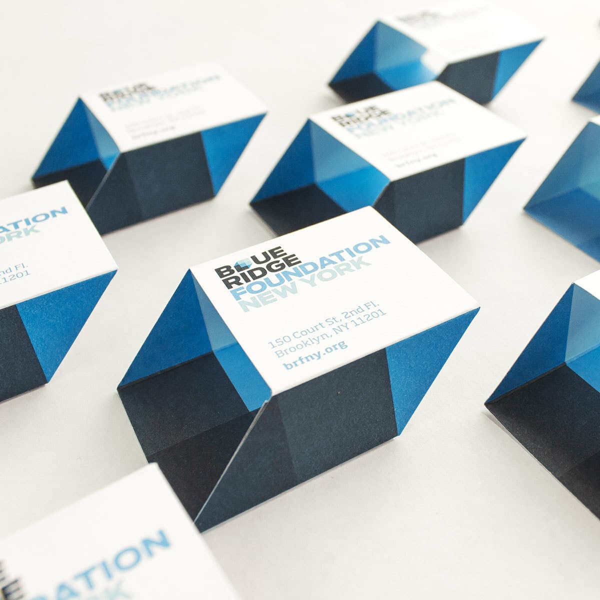 A set of uniquely shaped business cards for Blue Ridge Foundation New York are arranged on a white surface. The cards have a 3D geometric design with blue and dark blue sections. The text includes the organization's name, address, and website.