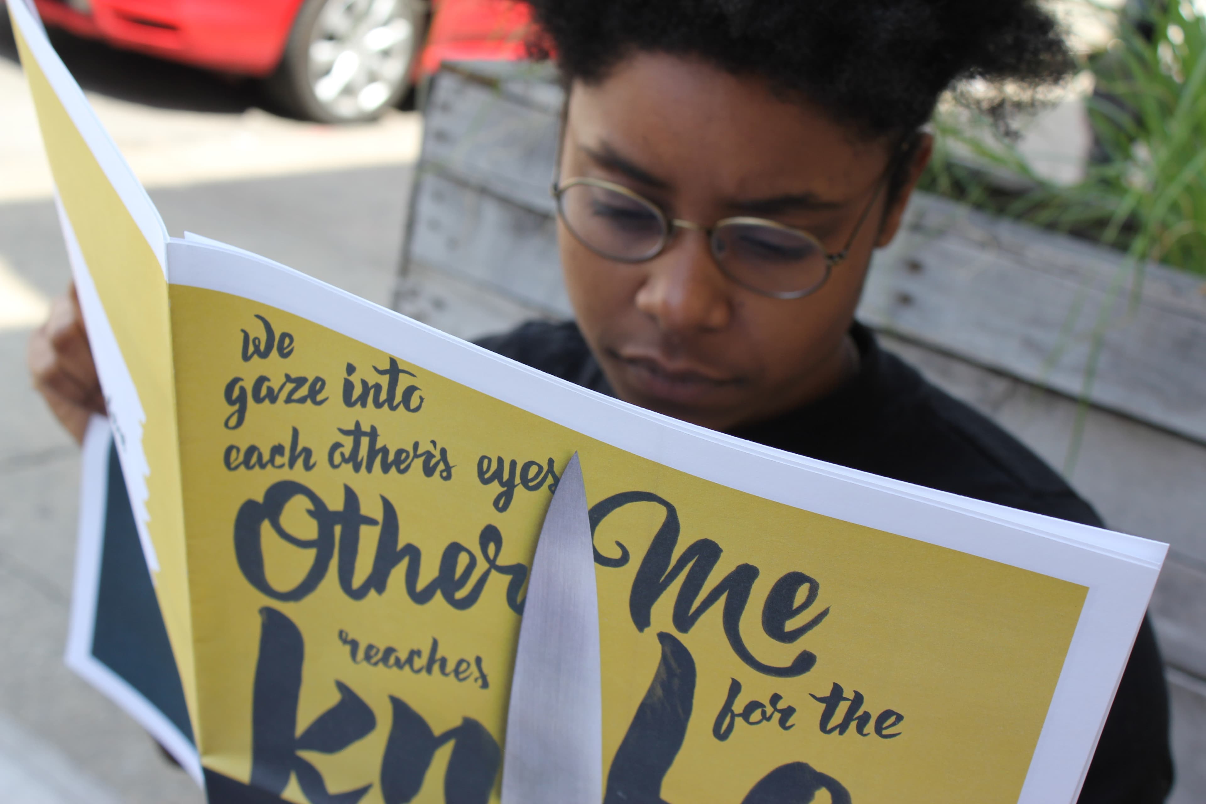 A person with glasses and dark curly hair intently reads a brightly colored publication. The visible text on the publication reads: "We gaze into each other's eyes" and "Me for the" in a stylized font. A red car is parked in the background.