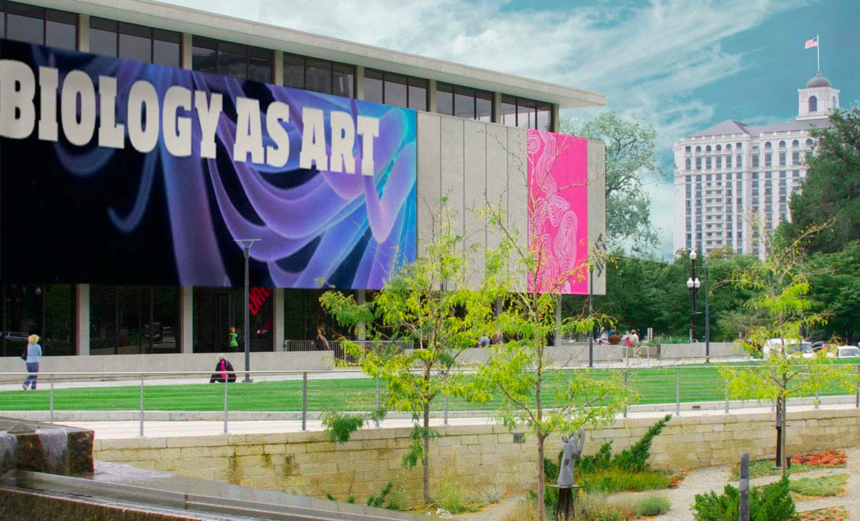 The image shows a modern building with large banners on its facade, one reading "BIOLOGY AS ART." The area in front has a landscaped garden with trimmed grass, trees, and a fountain. In the background, a tall historic building is visible against a blue sky.