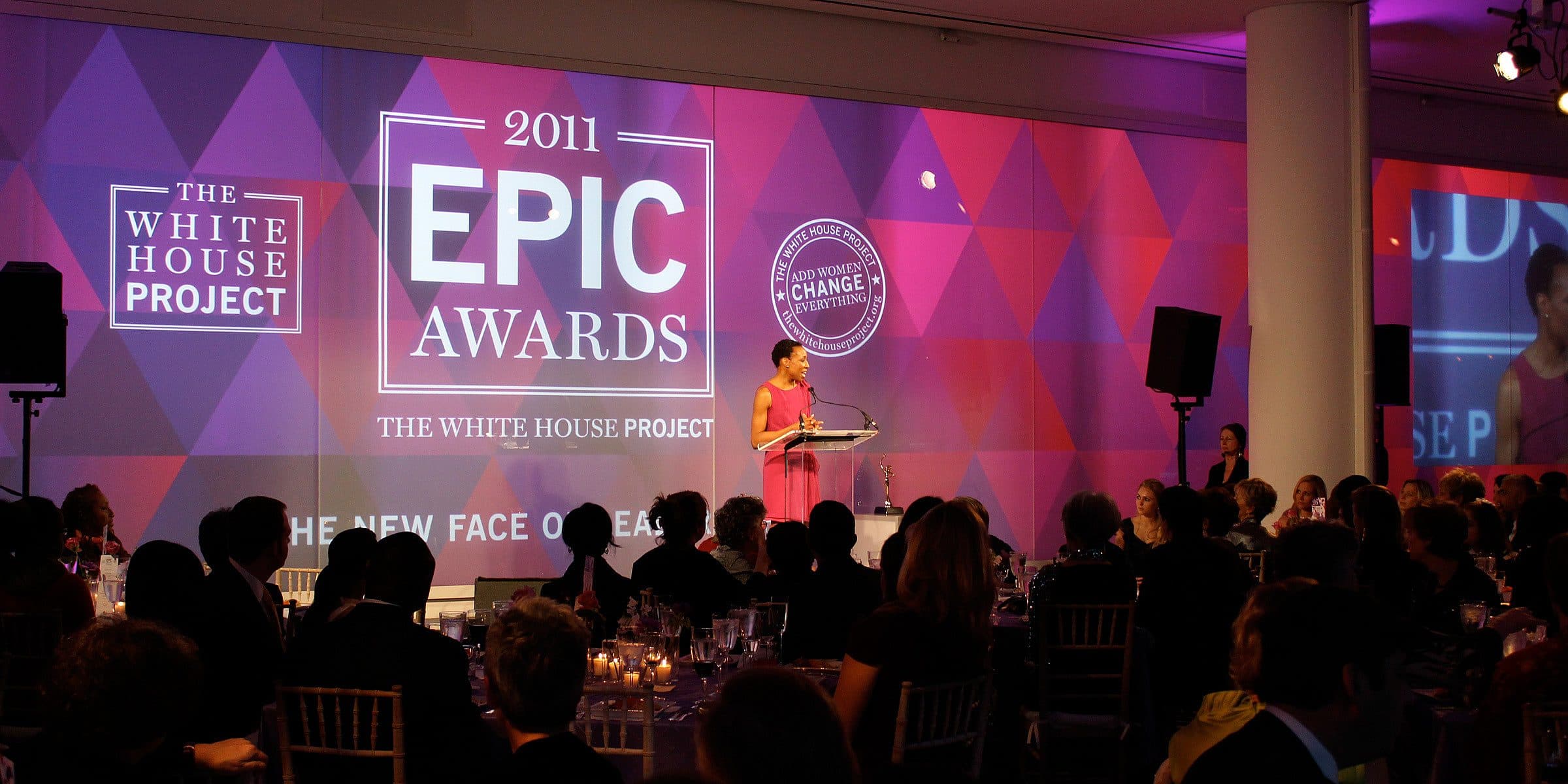 A speaker stands at a podium on a stage at the 2011 EPIC Awards hosted by The White House Project. The audience is seated at round tables, and a backdrop features the event's logos and theme, "The New Face of Leadership." The room is dimly lit with a purple hue.