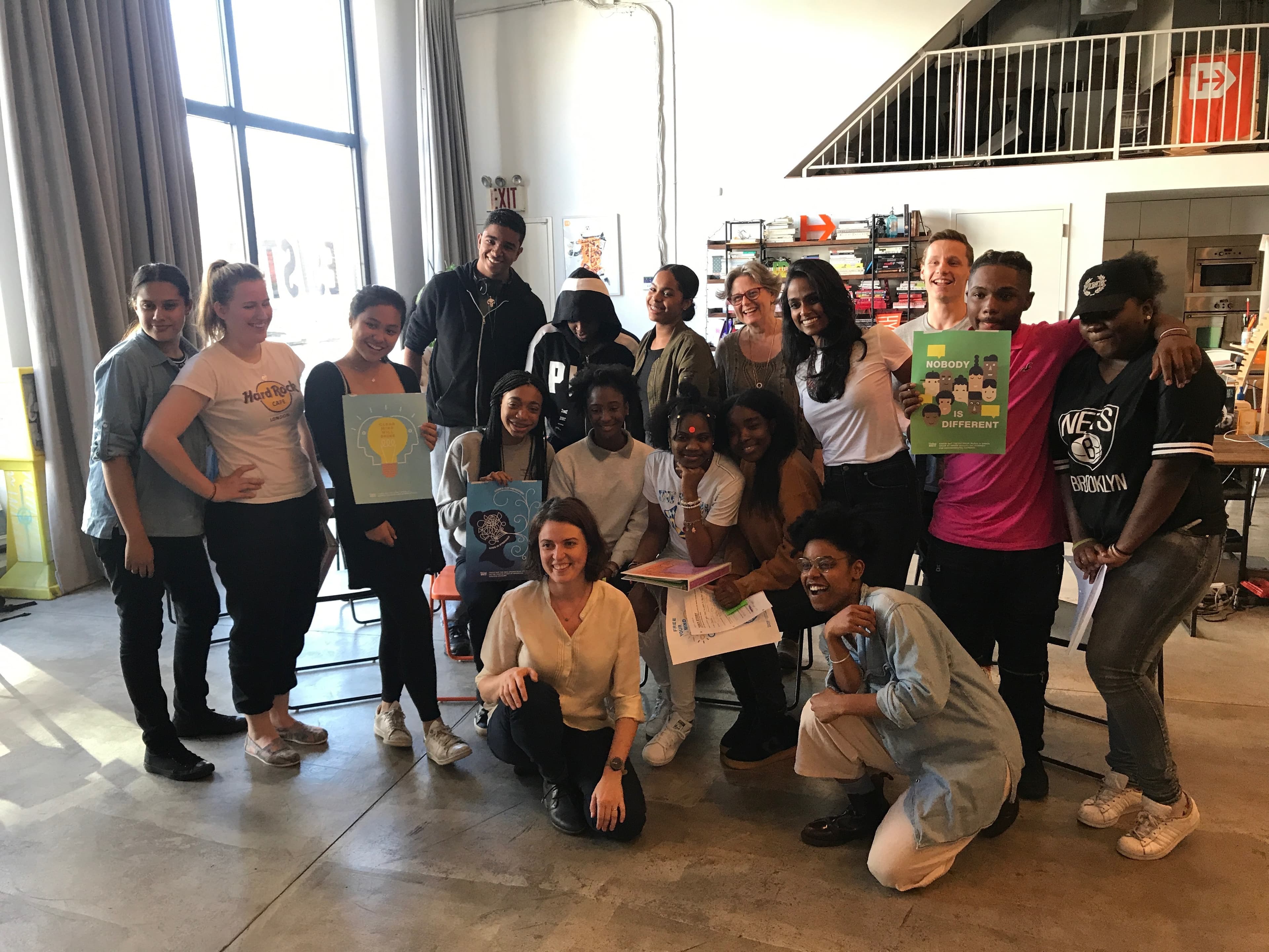 A diverse group of 16 people, including men and women, pose together in a well-lit indoor space. Some are holding colorful signs with messages like "Be Different." They appear happy and relaxed, with smiles and casual poses.
