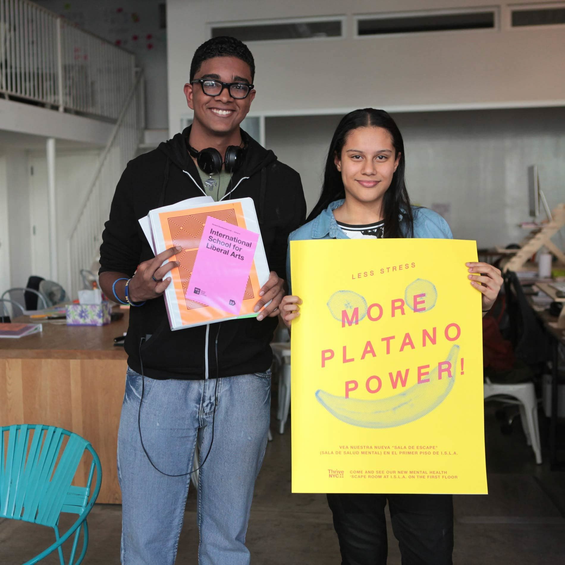 Two smiling students stand indoors in a bright, workstation-filled space. The taller student on the left holds two books titled "International School for Liberal Arts" and "World Literature." The student on the right holds a vibrant yellow poster that reads "More Platano Power!.