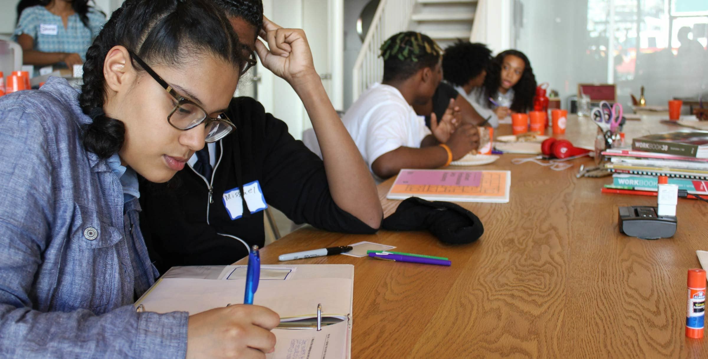 Several young people sit around a rectangular wooden table, engaging in group activities. In the foreground, a girl with glasses writes in a notebook, holding a pen. Others are engaged in conversation, and various office supplies, books, and papers are scattered on the table.