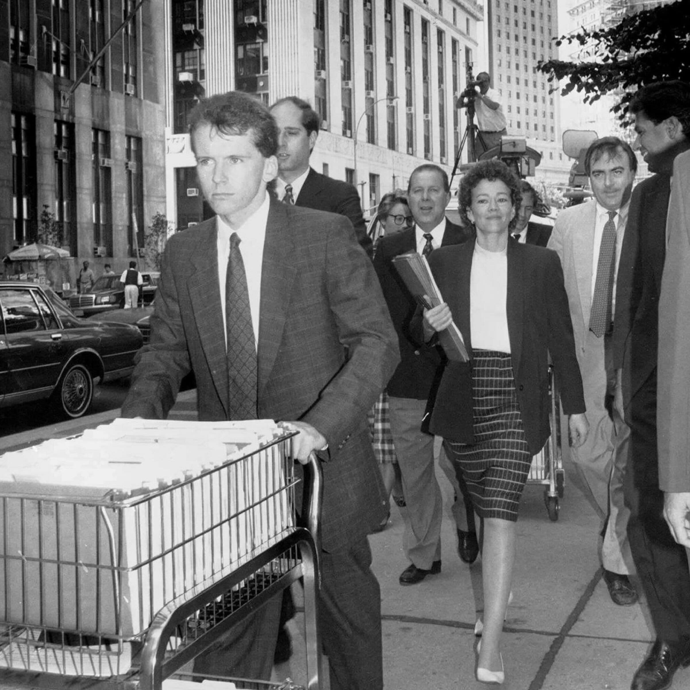 A black and white photo showing a group of people walking on a city street. In the foreground, a man pushes a cart filled with files. Behind him, a woman in a plaid skirt and blazer walks while holding a folder. Office buildings surround the group.