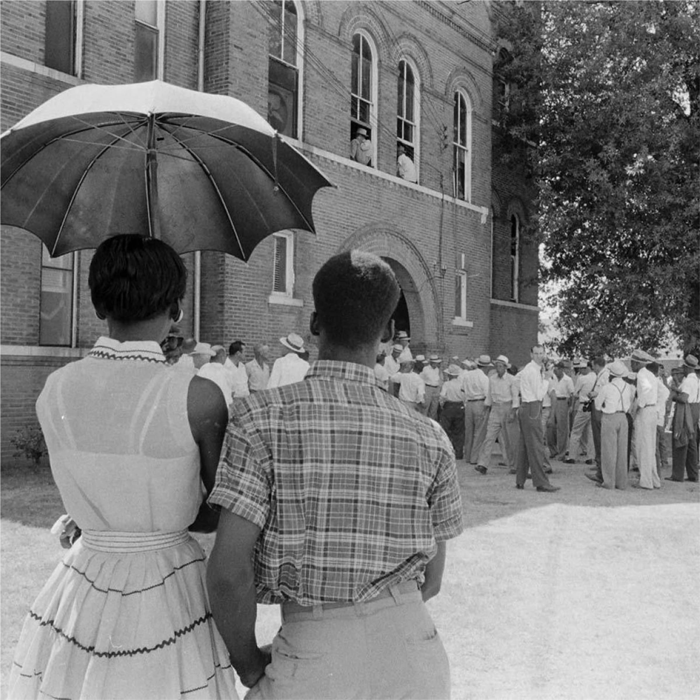 A Black woman and a Black man stand under an umbrella, facing away from the camera, observing a crowd gathered outside a brick building. Some people are seen looking out from the building's windows. The scene appears to be from the mid-20th century.