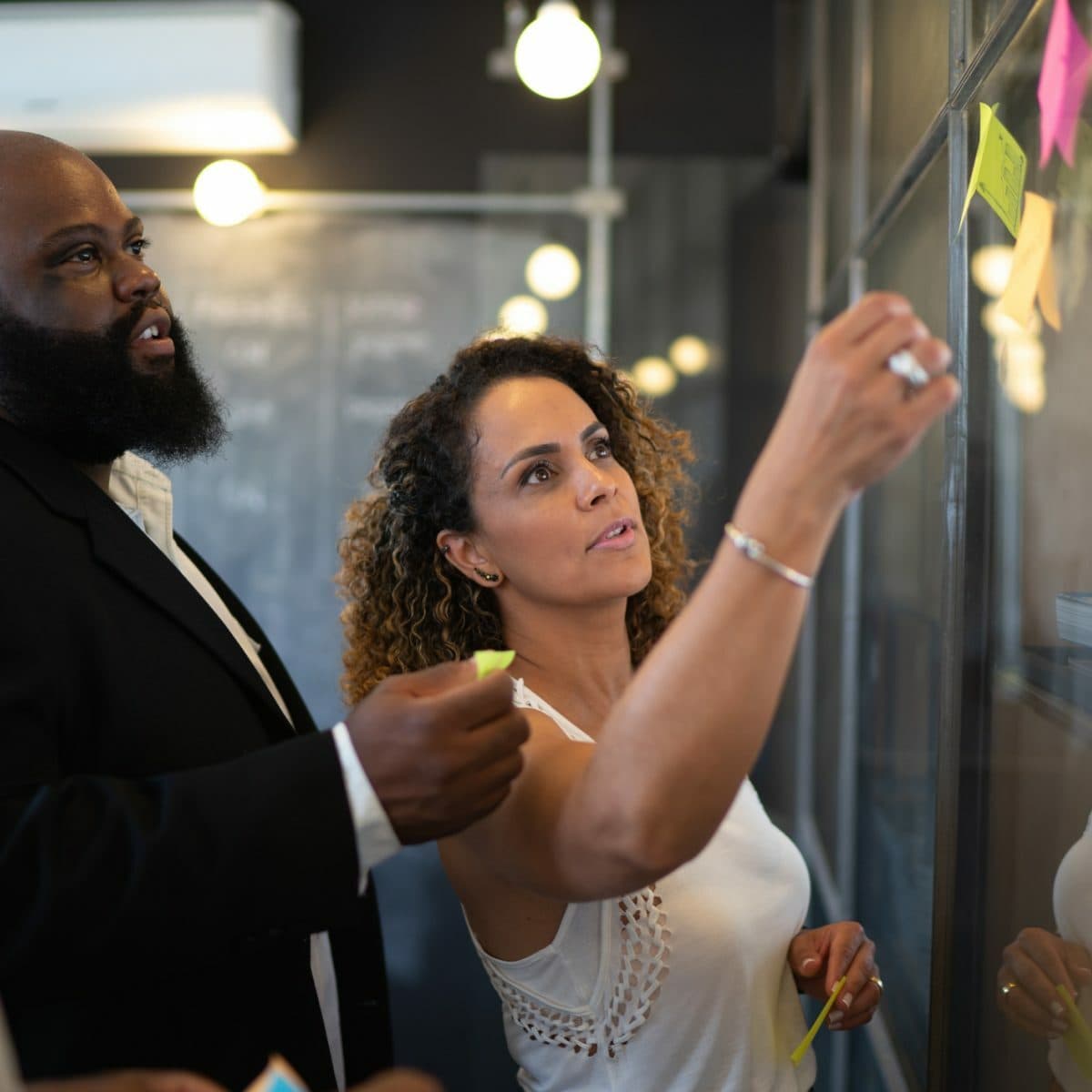 Two colleagues collaborate by placing sticky notes on a glass wall in an office. The man, in a dark suit, observes while the woman, in a white sleeveless top, focuses on sticking a note. Both appear engaged in brainstorming or project planning.