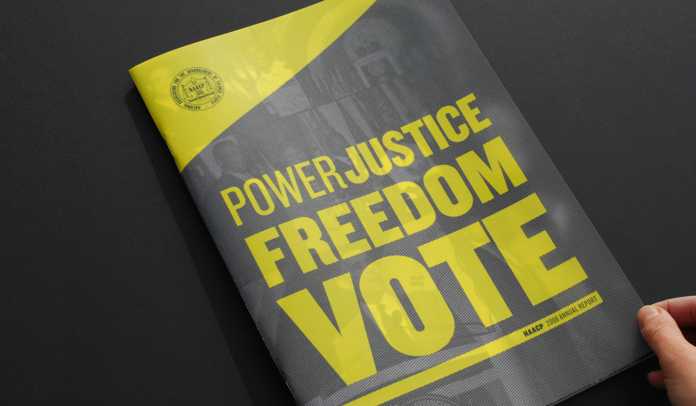 A hand holding a magazine with a black and yellow cover. The magazine features bold, large text in yellow and white that reads "POWER, JUSTICE, FREEDOM, VOTE" and "NAACP 2022 ANNUAL REPORT" in smaller print. The background includes a subtle image of people.