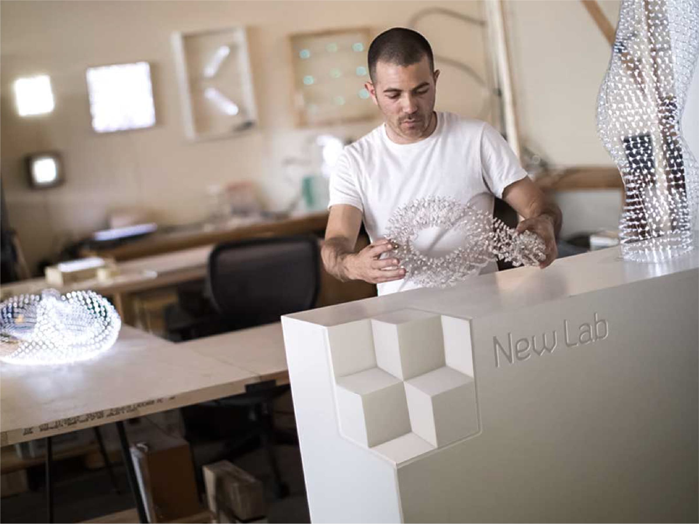A person wearing a white t-shirt stands behind a counter labeled "New Lab," handling a geometric light sculpture. The room is filled with more light sculptures and various workshop tools, suggesting a creative and technical workspace.