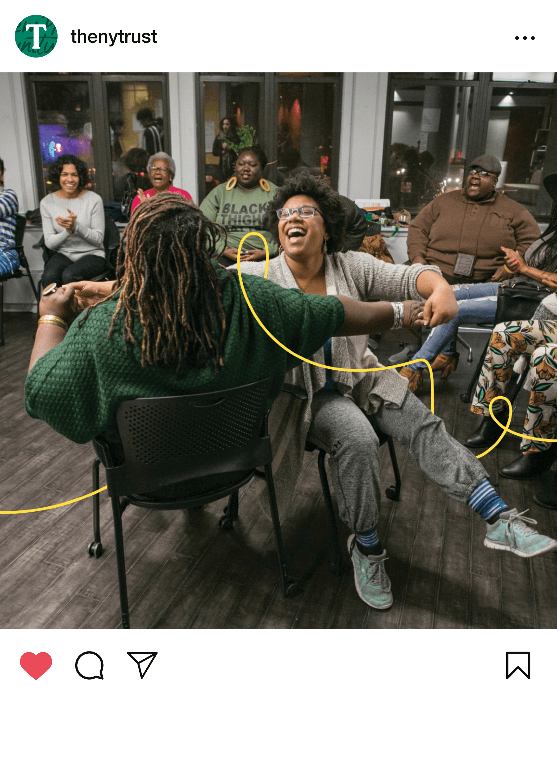 A lively group of diverse adults is gathered in a room, smiling and laughing. Two women in the front are playfully entangled in yellow string, creating a joyful and lighthearted atmosphere. Some people are seated, while others engage with the activity in the background.