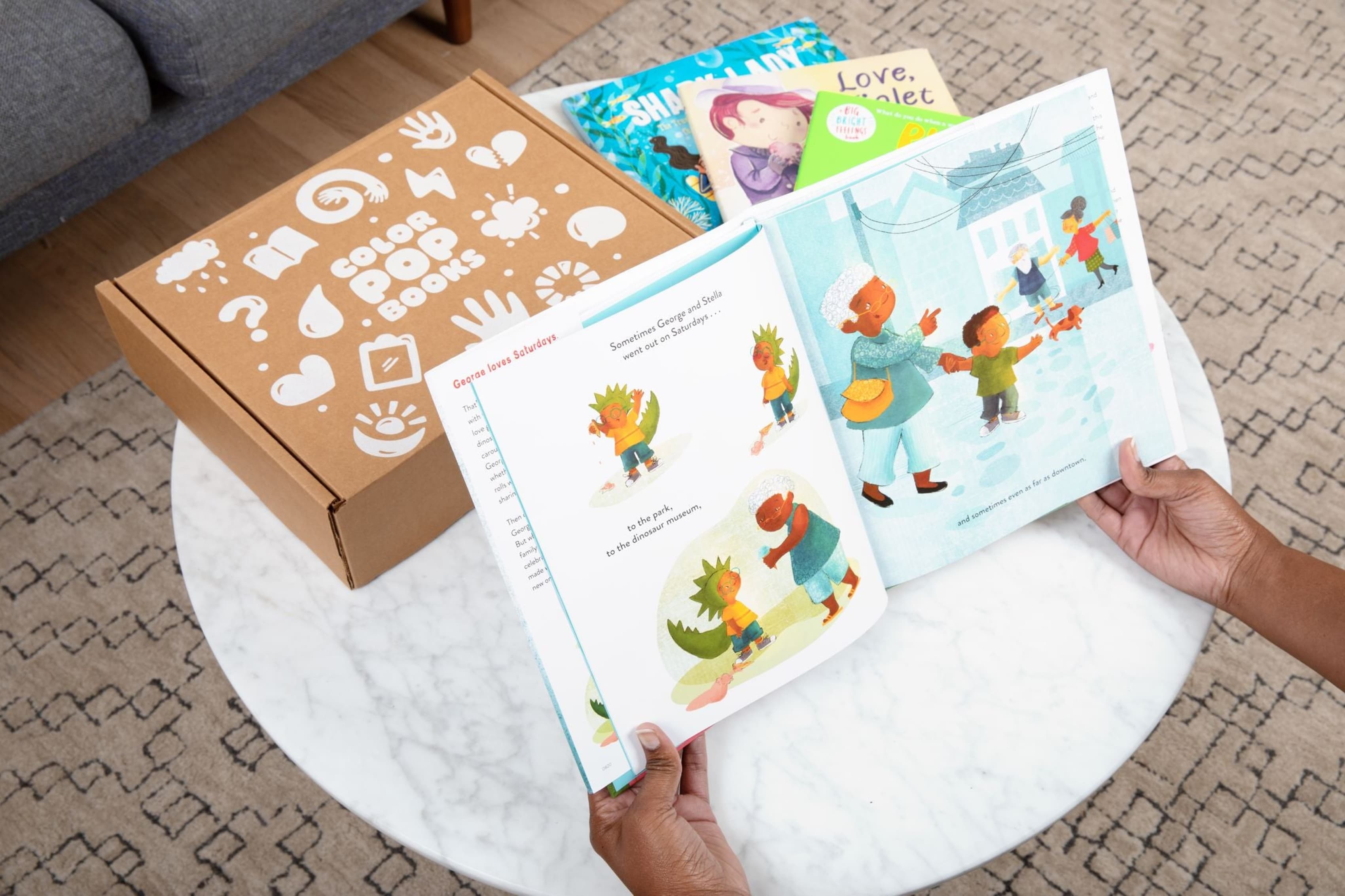 Hands holding open a children's book with colorful illustrations on a round, white marble table. Beside the book is a brown box with white graphics labeled "COCO BOX BOOKS," and several other children's books with vibrant covers are resting on the table.