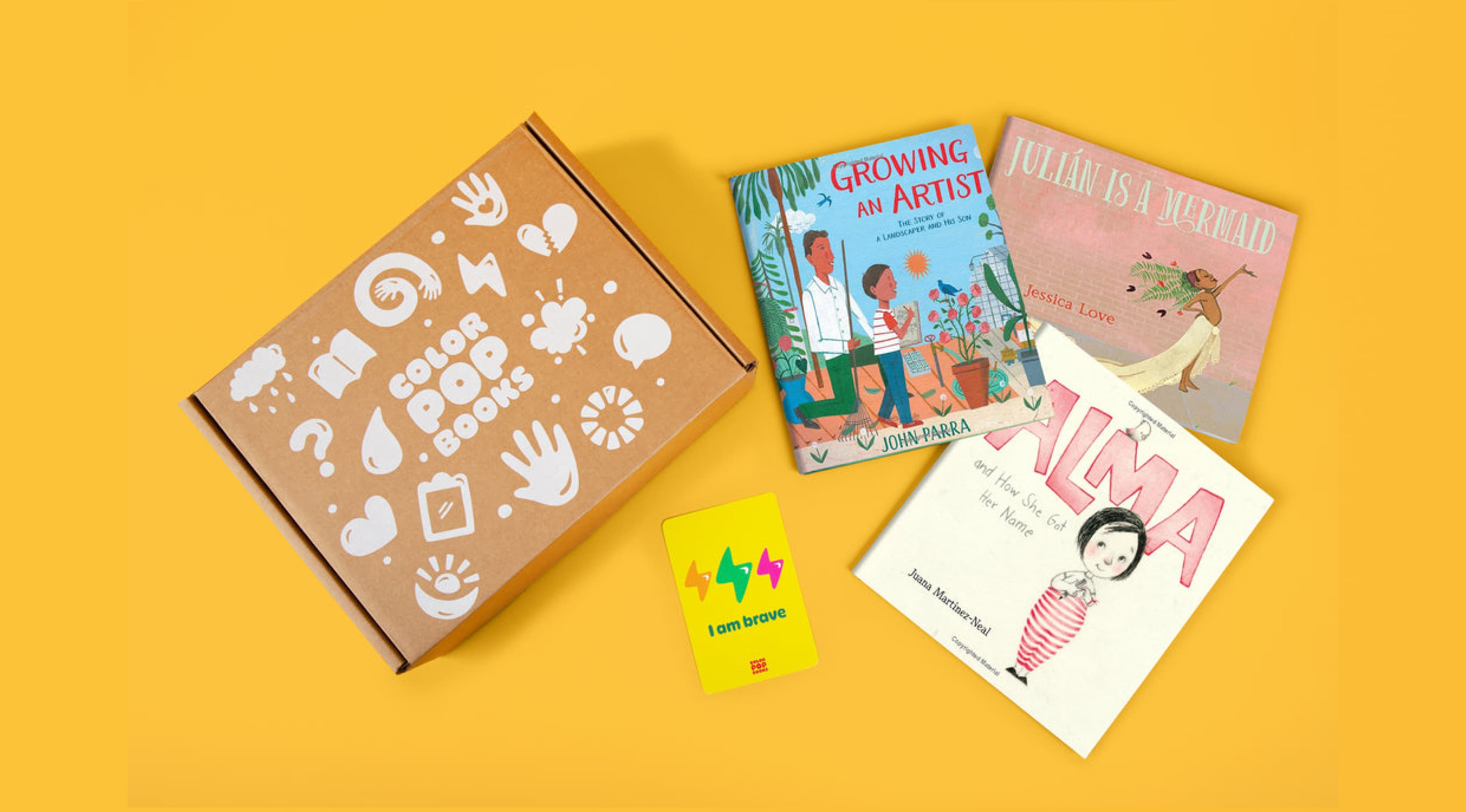 A Color Pop Books box is open on a yellow surface, revealing three children's books: "Growing an Artist," "Julian is a Mermaid," and "Alma." Additionally, there is a card with colorful bolts and the words "I am brave.