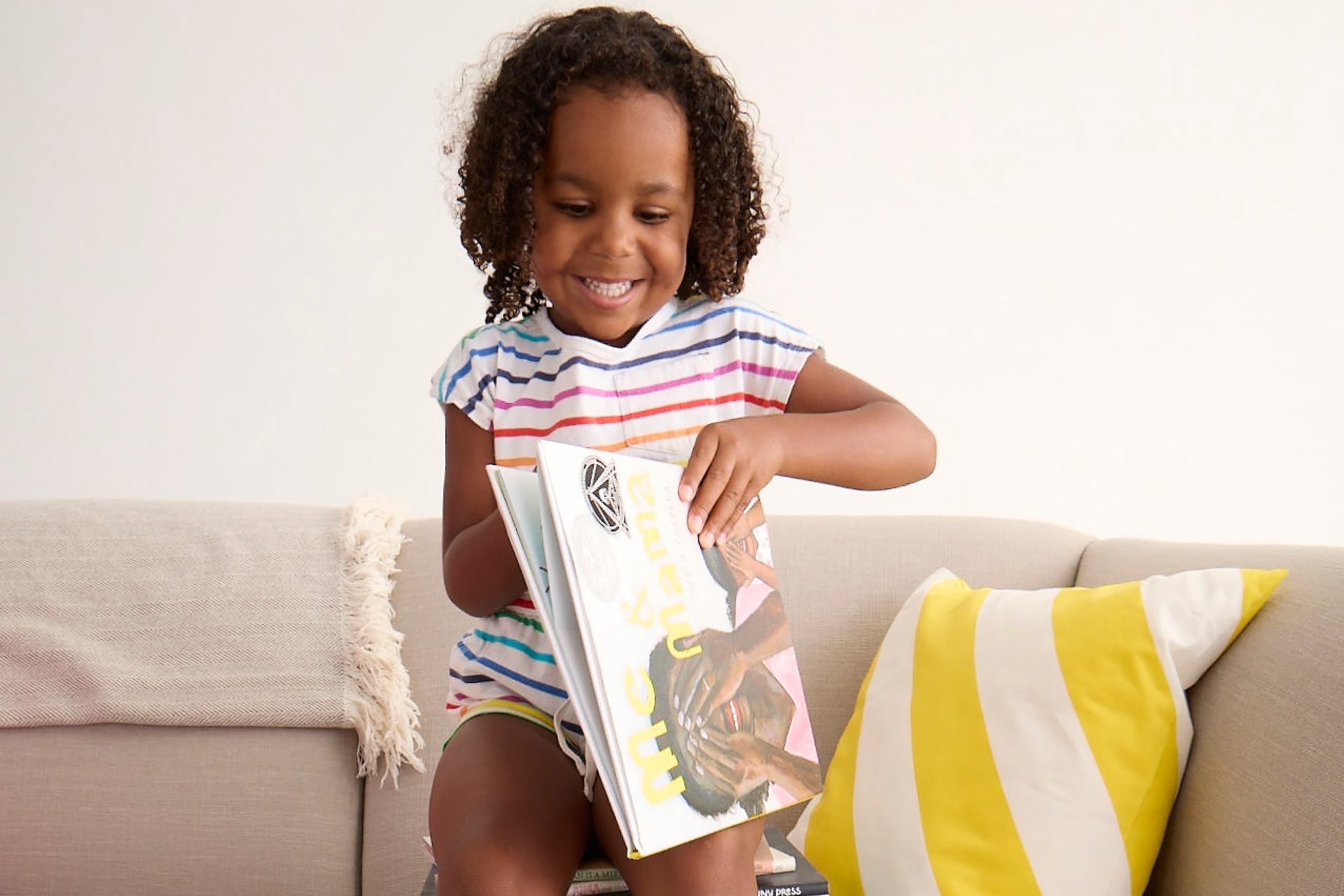 A young child with curly hair, wearing a colorful striped shirt, sits on a beige couch with yellow and white pillows, smiling and reading a large book. The setting is bright and cheerful.