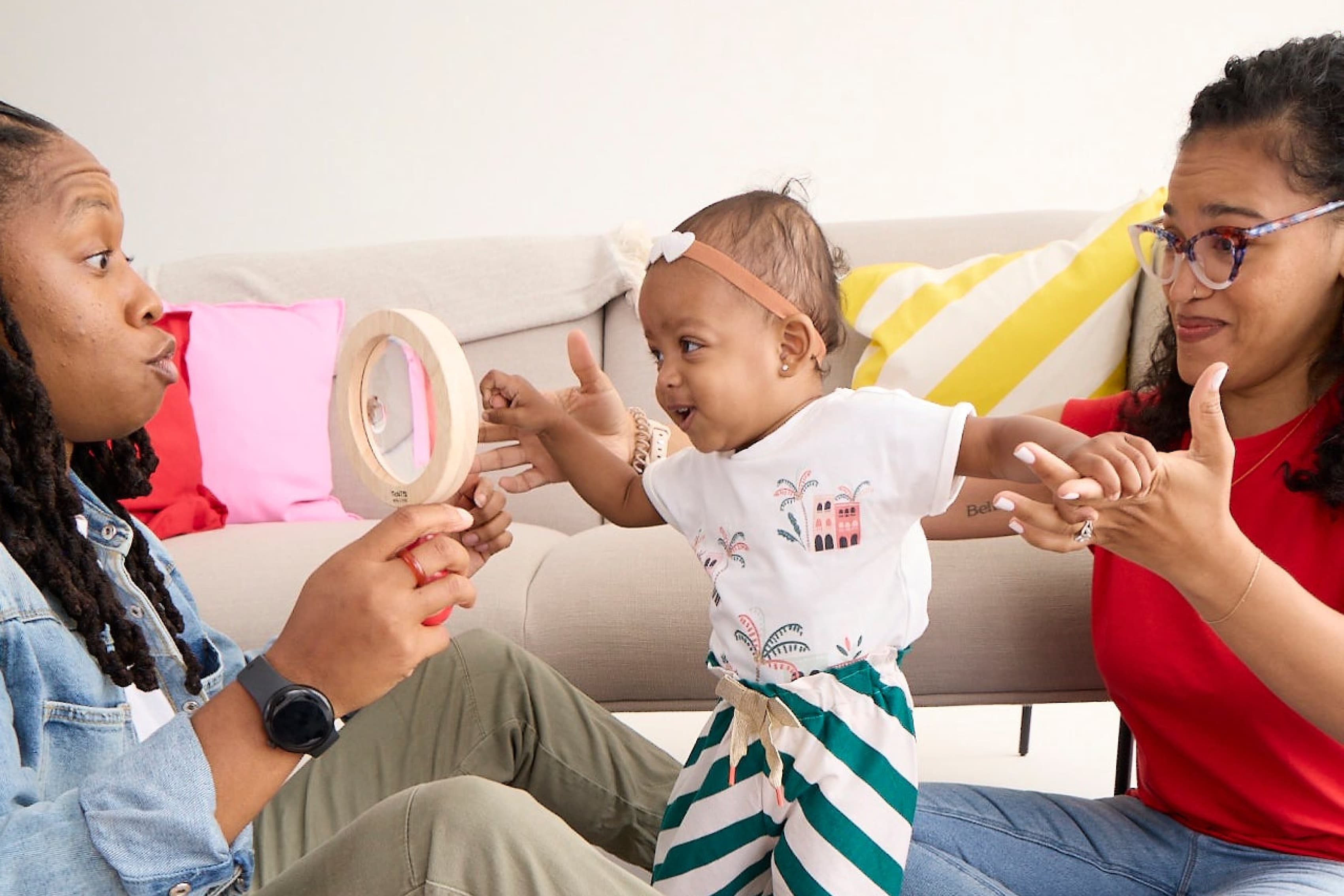 Two adults interact with a small child who is standing. One adult, seated on the floor, holds a mirror for the child, who is reaching towards it and smiling. The second adult, wearing glasses, sits on the couch and smiles, extending their hands towards the child.