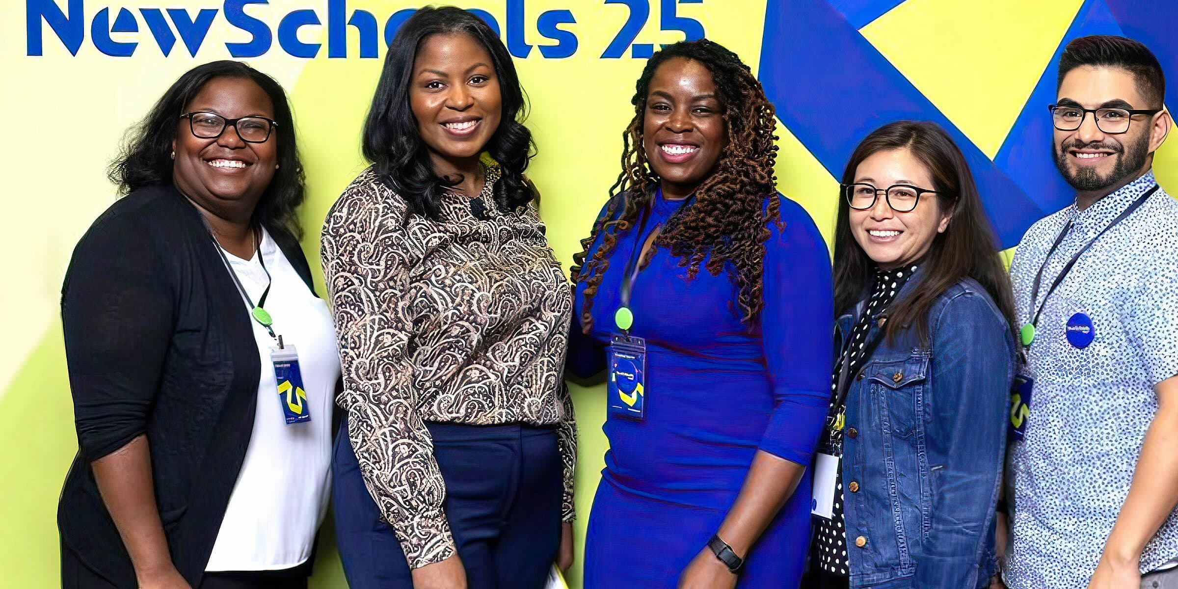 A group of five people smiling and posing in front of a wall with a colorful graphic and the text "NewSchools 25" written on it. They are wearing business casual attire and name tags. The background features a yellow star on a blue zigzag design.