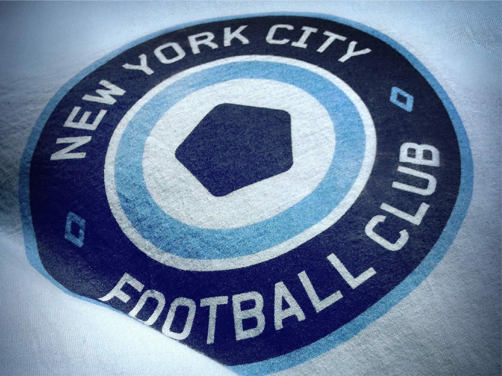 Close-up image of the New York City Football Club logo printed on fabric. The logo features concentric blue circles with the club's name in white text surrounding a light blue hexagon in the center.
