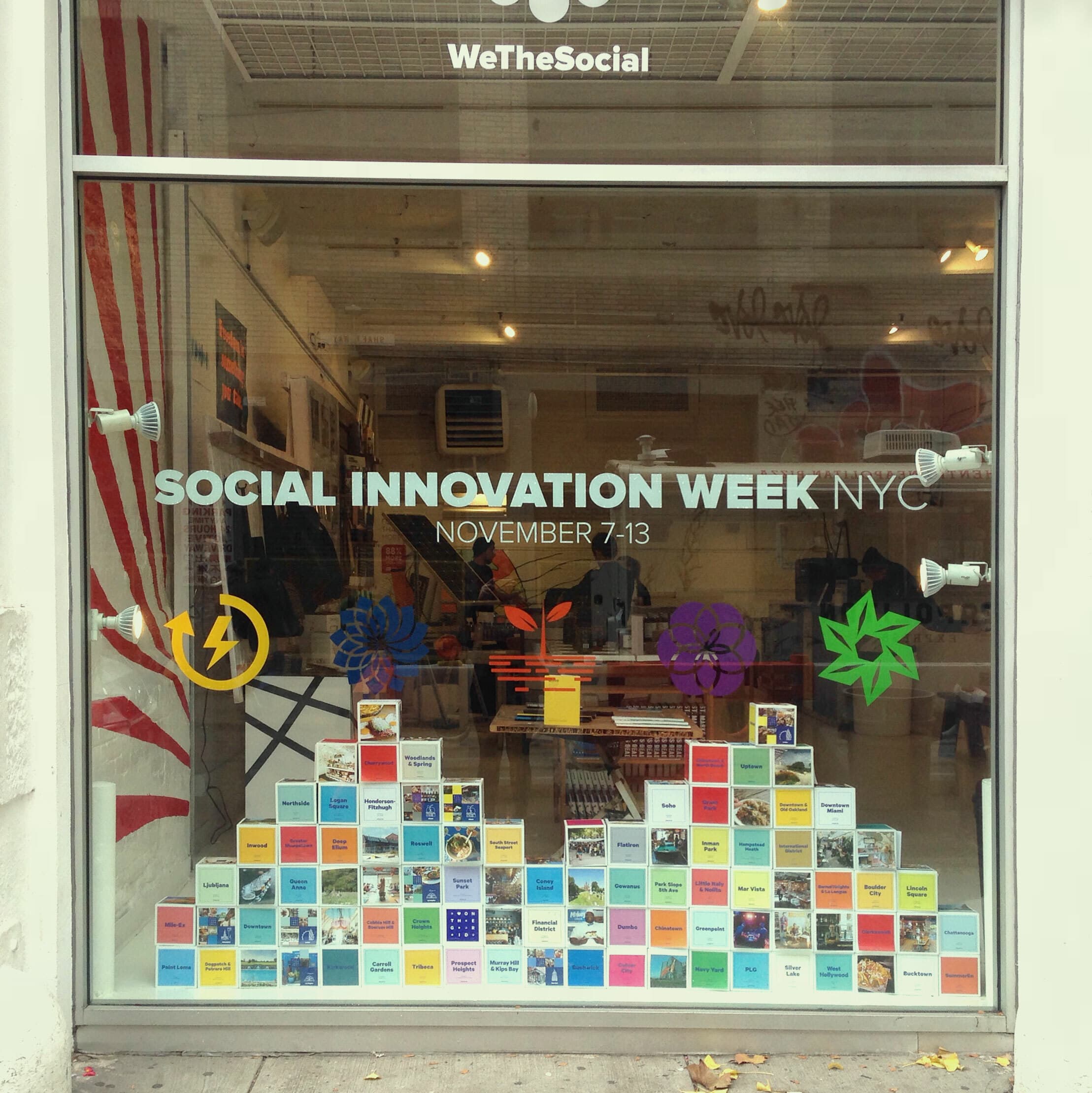 Window display promoting "Social Innovation Week NYC" taking place from November 7-13. The display includes colourful post-it notes, vivid graphics including flowers, and photos arranged on the glass. Inside, there are more displays and a spacious, well-lit interior.