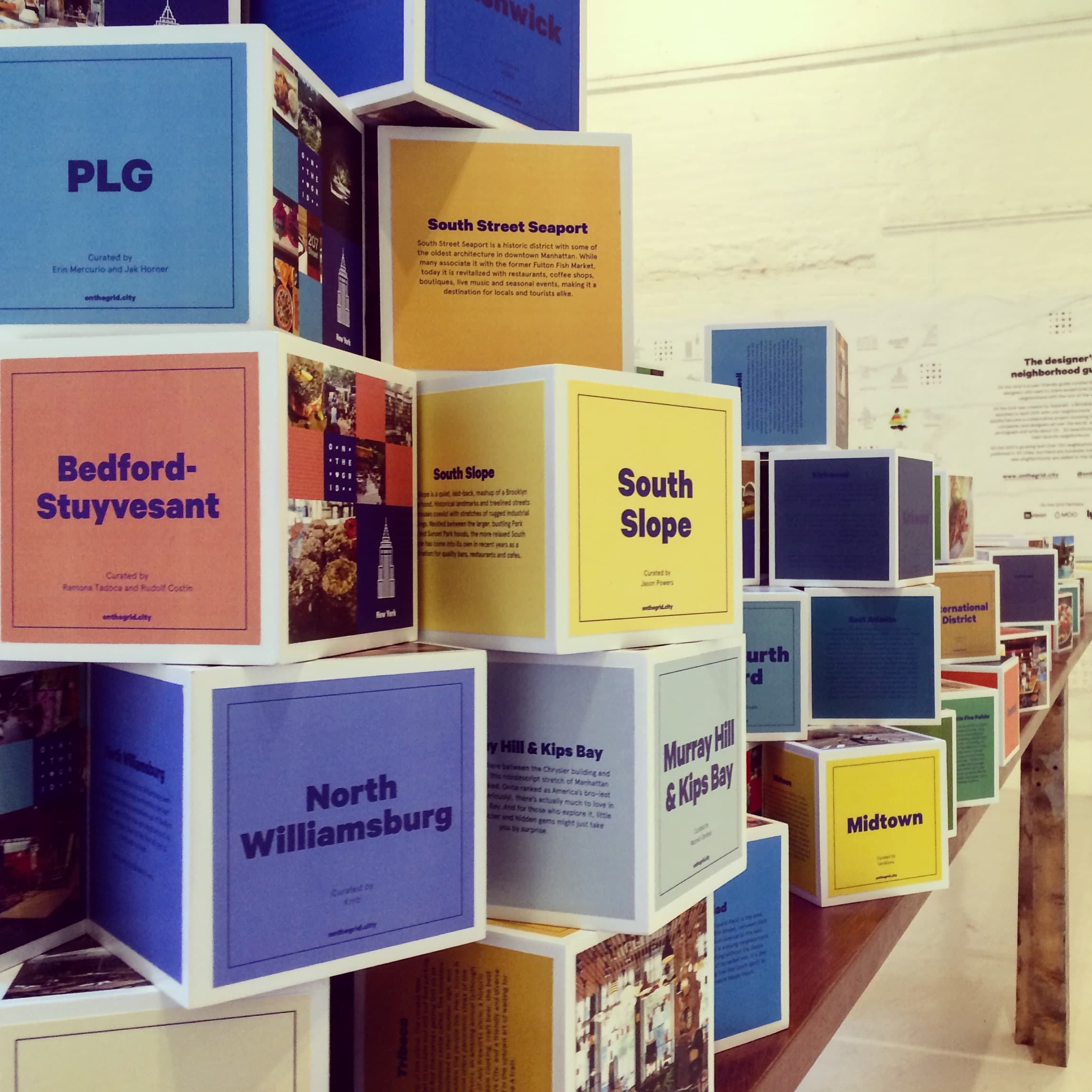 Stacks of colorful blocks on a table, each labeled with names of various New York City neighborhoods such as Bedford-Stuyvesant, North Williamsburg, South Slope, and Midtown. The blocks also feature descriptions and small images related to each neighborhood.