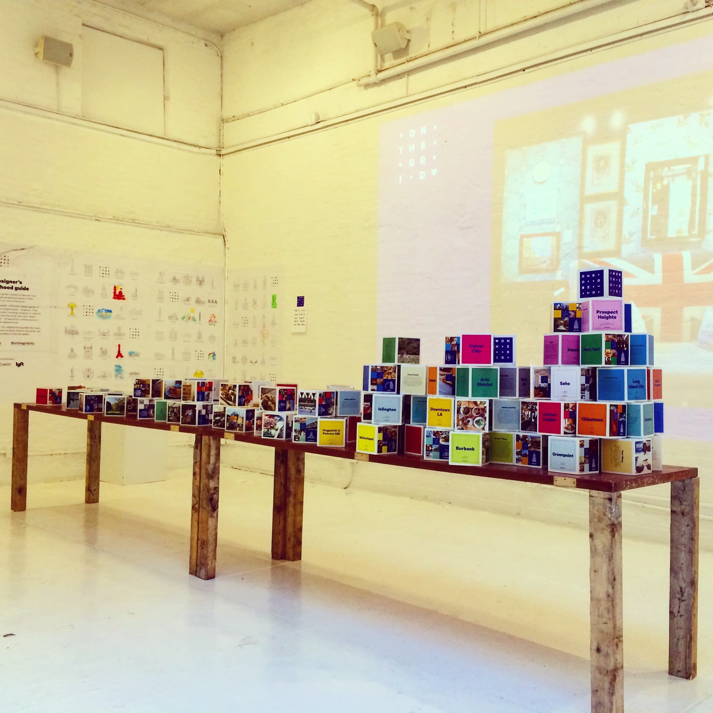 A modern art gallery features a rustic wooden table lined with colorful books arranged in a tiered, pyramidal structure. The white walls display various artworks, and a large projection of artwork is visible on one wall. The space is bright and minimalistic.