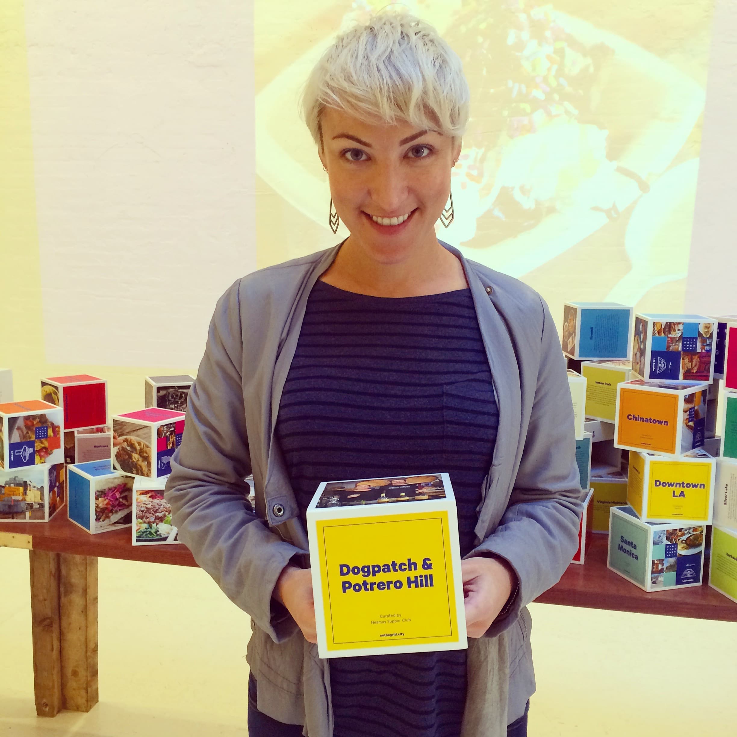 A person with short, light-colored hair and earrings stands indoors, smiling and holding a cube labeled "Dogpatch & Potrero Hill." They wear a gray jacket over a dark striped shirt. Colorful cubes with various labels are arranged on a table in the background.