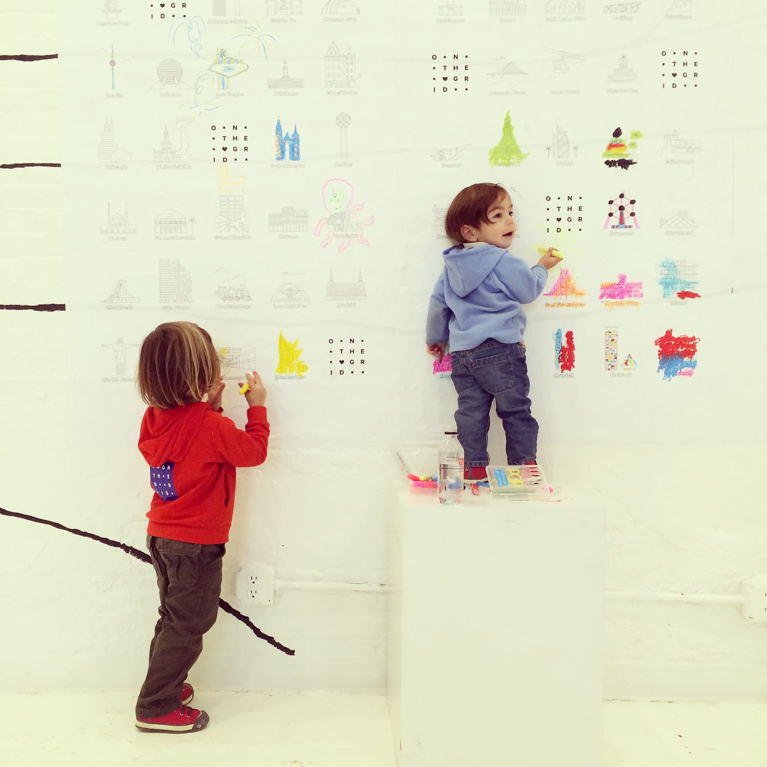 Two young children are drawing on a large white paper covering a wall. The child on the left, wearing a red hoodie, draws while standing on the floor. The child on the right, wearing a blue hoodie, draws while standing on a white pedestal. Colored drawings are visible on the paper.