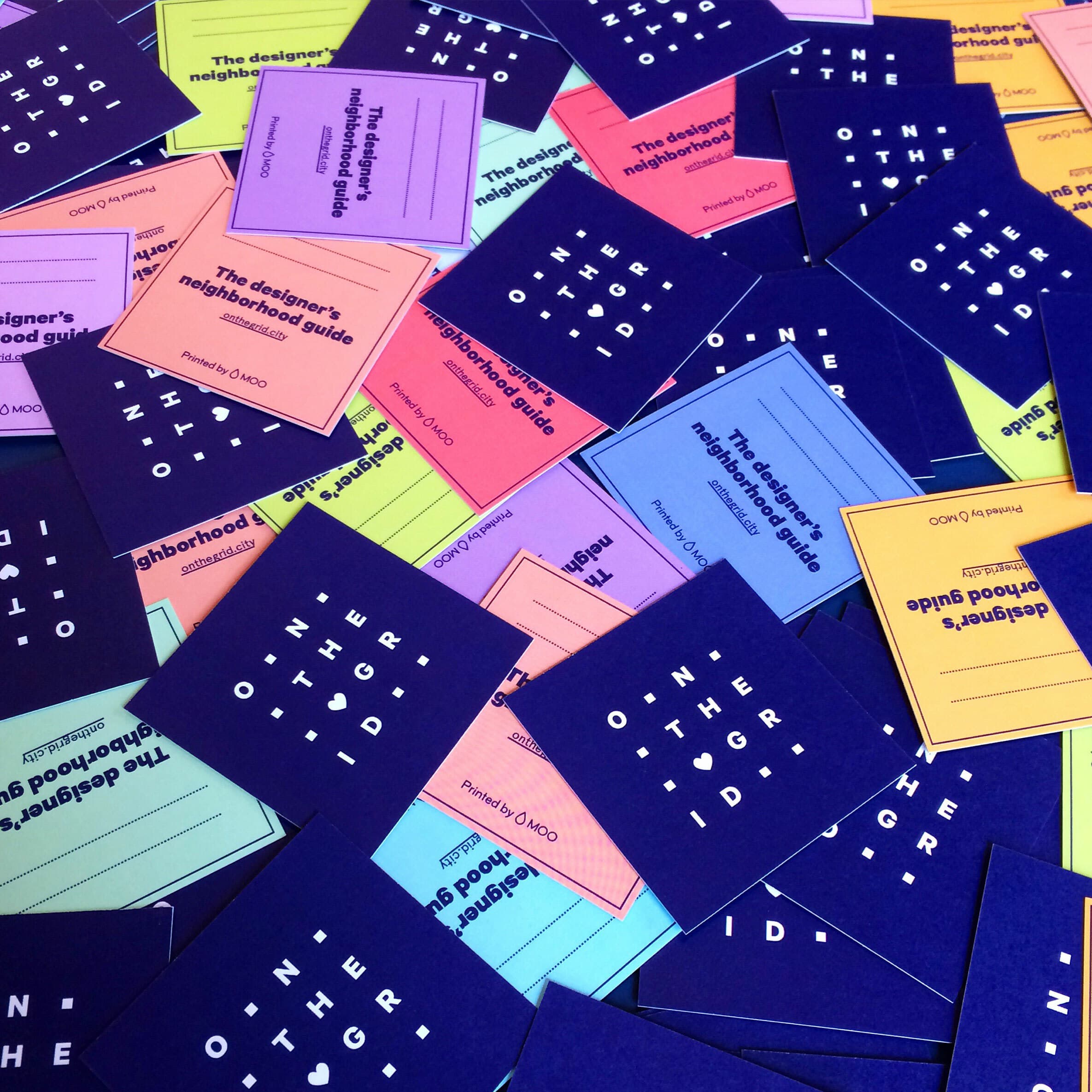 A vibrant display of colorful cards scattered on a surface. The cards are in shades of blue, pink, green, yellow, and orange, each labeled as "The designer's neighborhood guide" and "On the Grid," with small informative text printed on them.