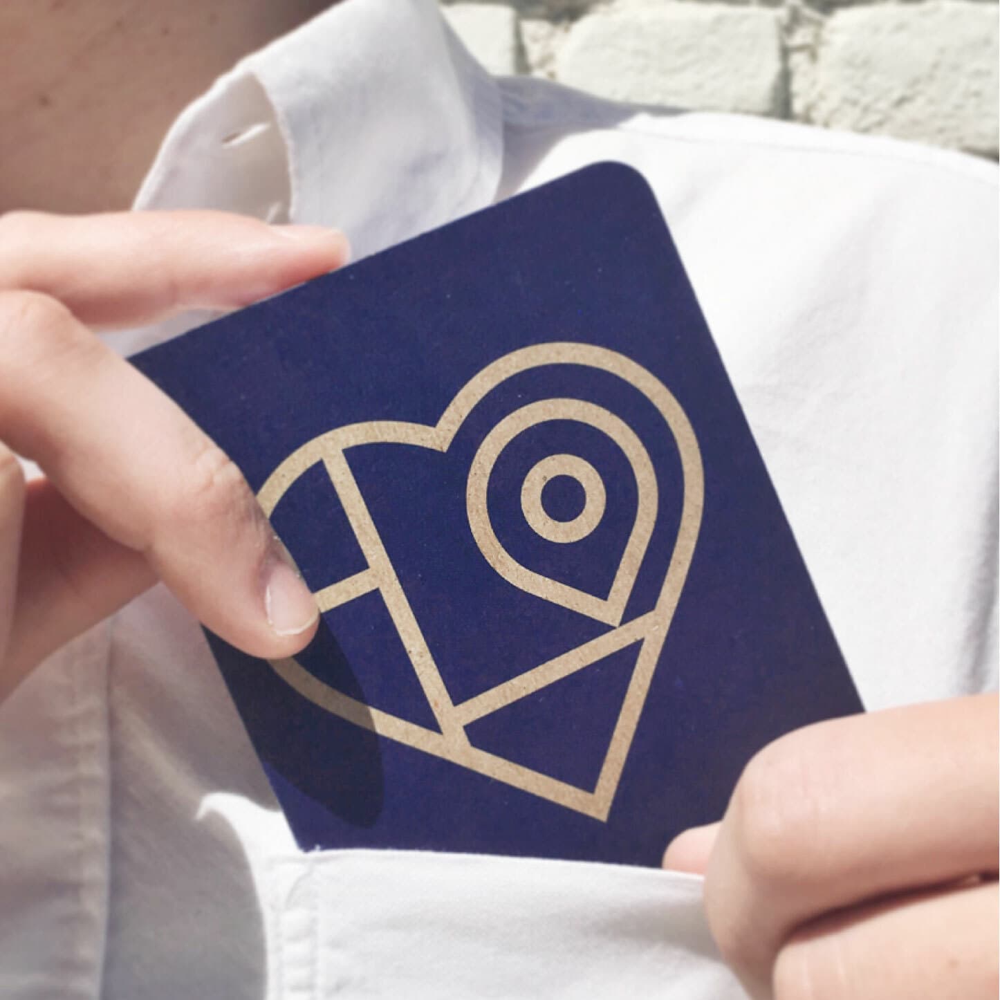 A person wearing a white shirt puts a dark blue card with a gold geometric heart design into their shirt pocket. The card features shapes including a circle and various triangles that form the heart. The background appears slightly blurred.