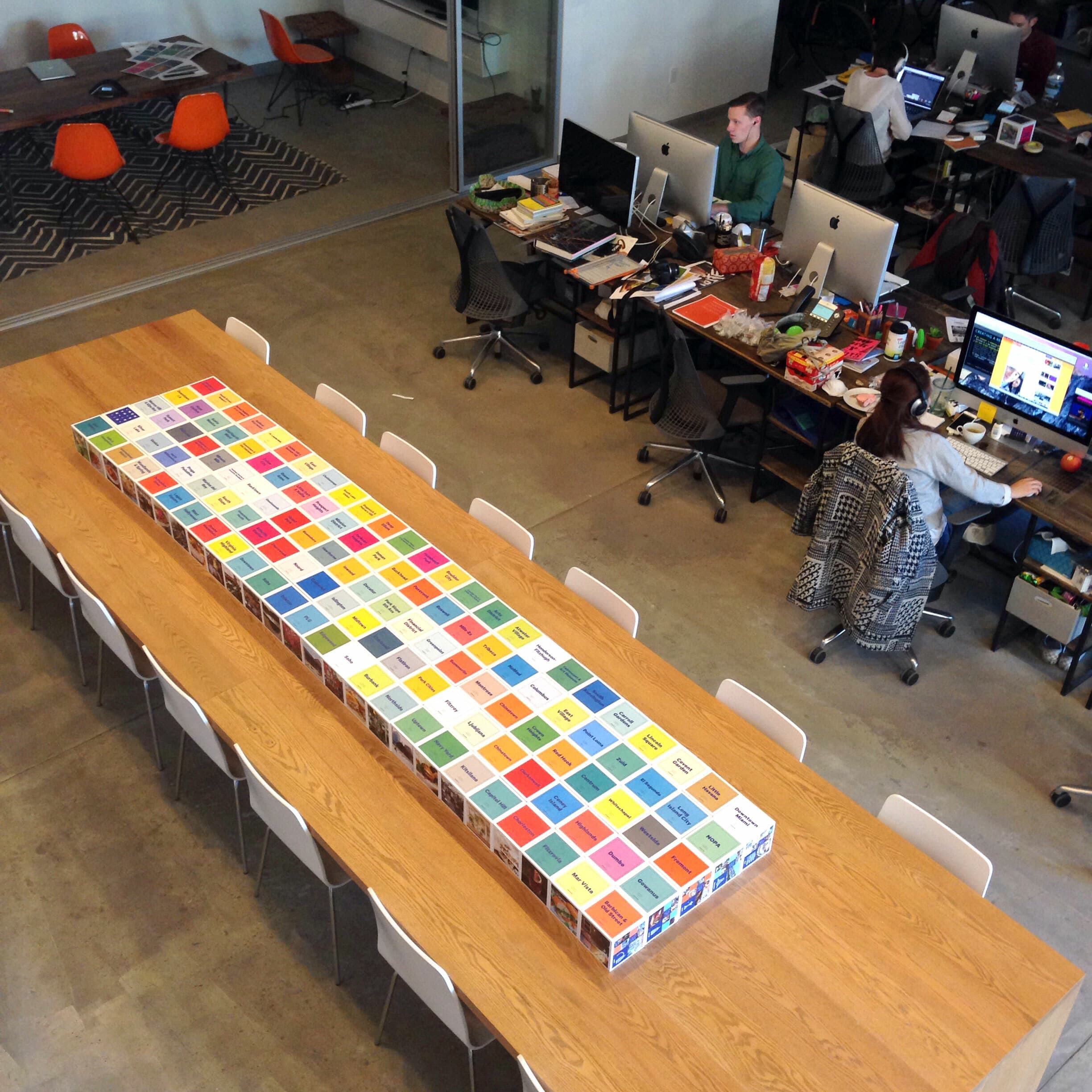 A long wooden table is fully covered with colorful boxes arranged in a grid pattern. The table is surrounded by white chairs. In the background, two people are working at a table with computer monitors, and an open space with chairs is visible.