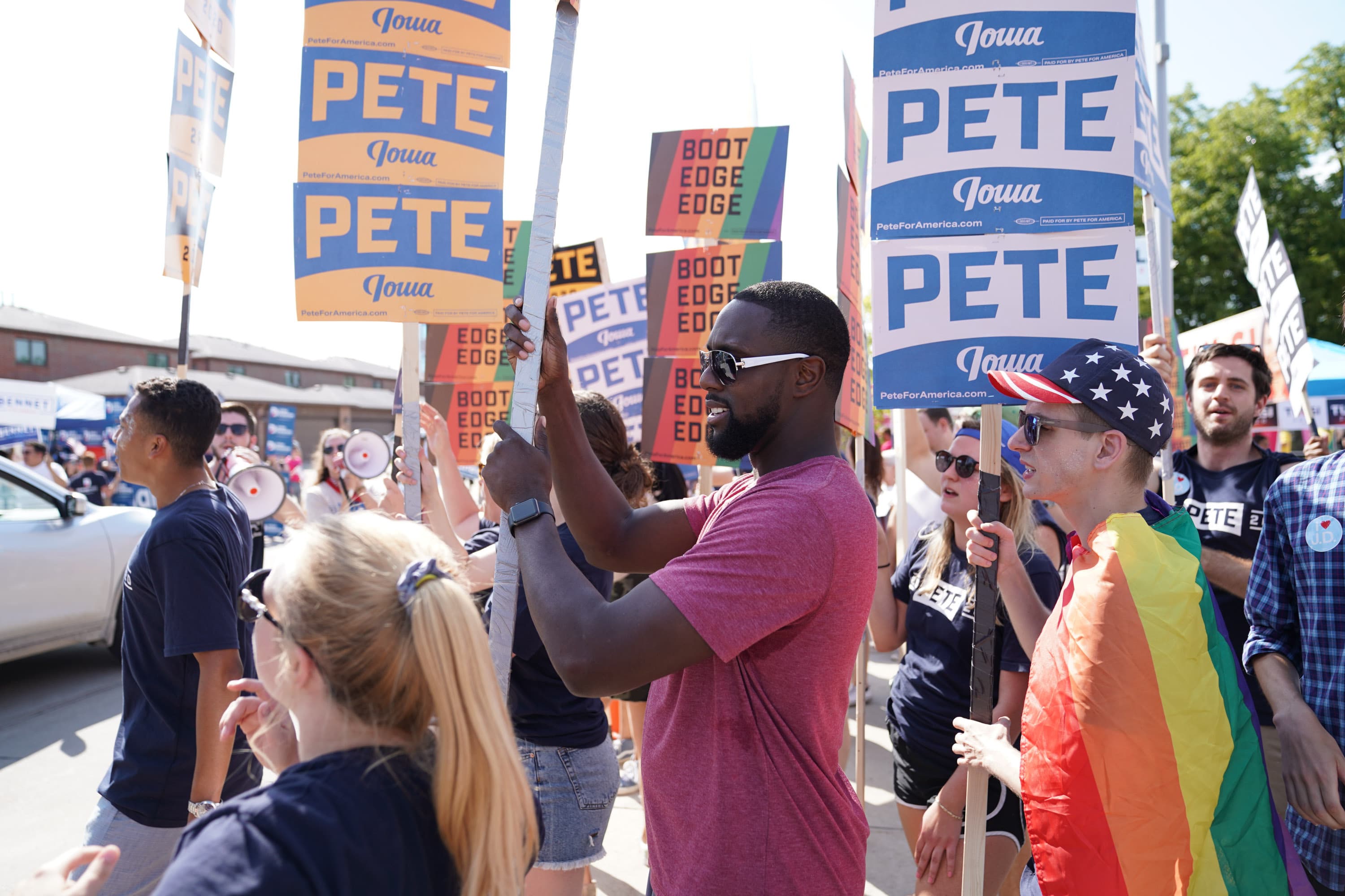 A group of people is holding signs that say "Iowa" and "Pete" at an outdoor event. One person is draped in a rainbow flag, and another person is wearing sunglasses and a red shirt. The background shows more people and trees.