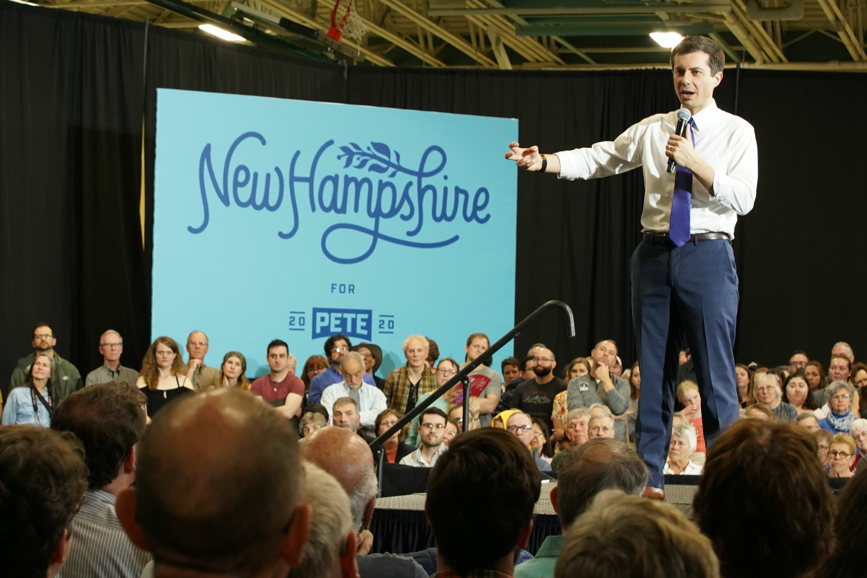 A person standing on stage in front of a "New Hampshire for Pete" sign, holding a microphone and gesturing while speaking to a large seated audience. The audience appears attentive and engaged. The setting seems to be an indoor venue with overhead lights.