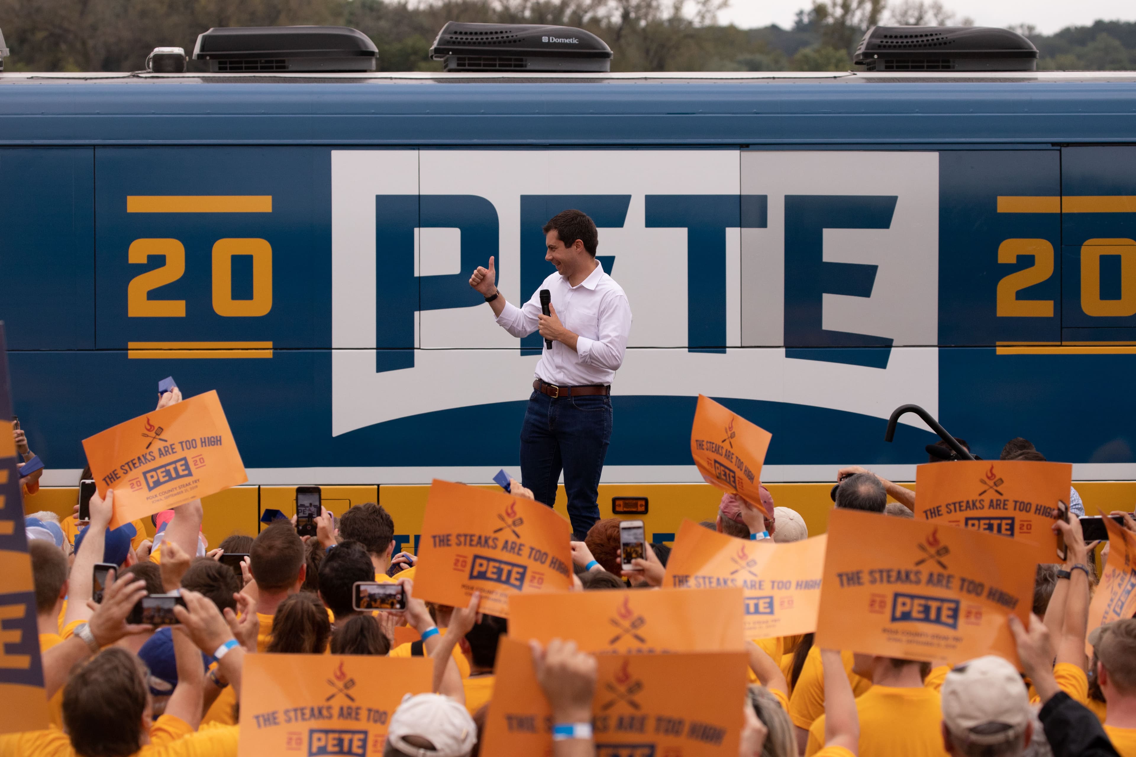 A person in a white shirt giving a thumbs up stands on a stage in front of a blue and yellow campaign bus with the name "Pete" on it. The crowd around the stage holds orange signs that read "THE STEAKS ARE TOO HIGH.