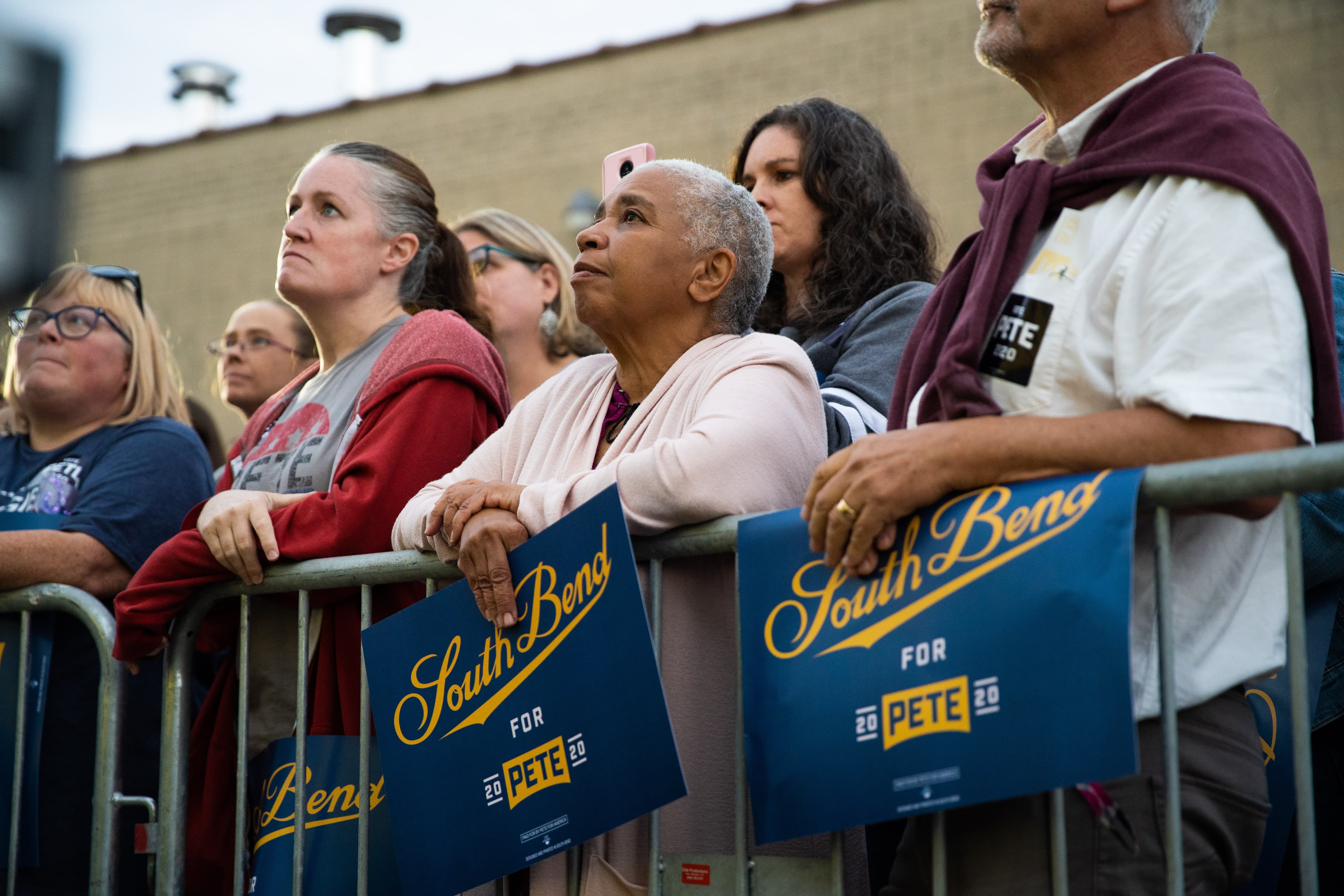 A diverse group of people stand closely together behind a metal barricade, attentively listening at an outdoor event. Some hold blue signs that read "South Bend for Pete 2020." The background shows part of a building.