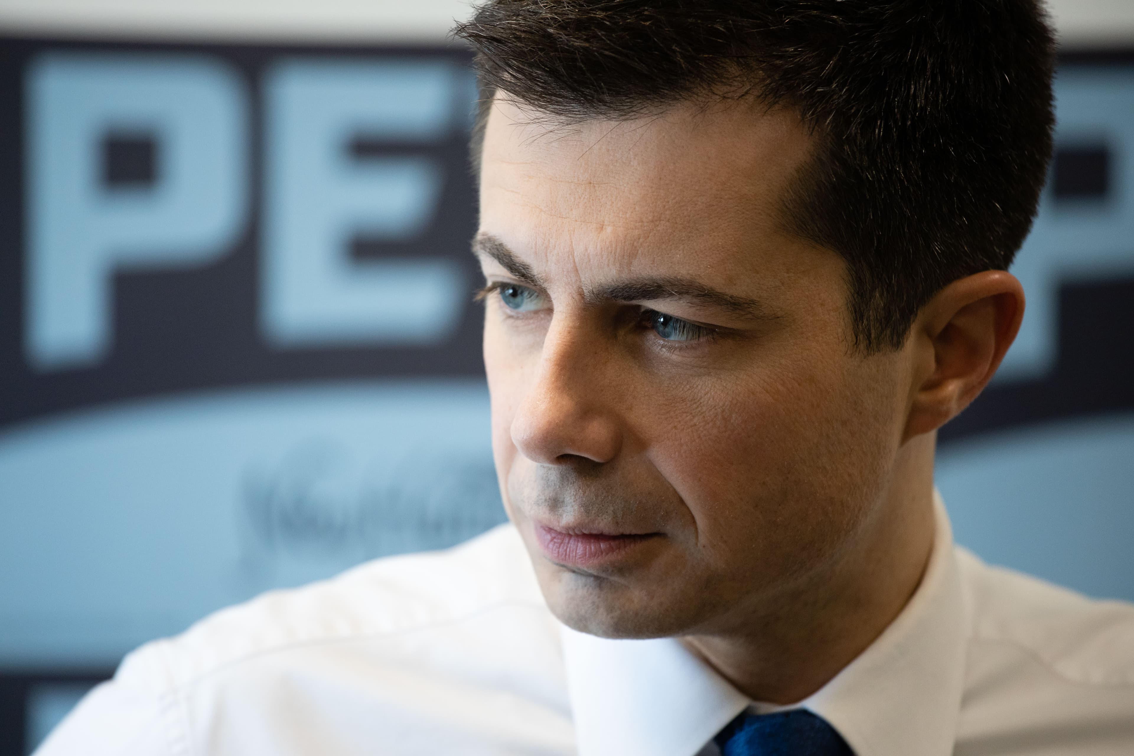 Close-up of a man with short dark hair, wearing a white shirt and blue tie, looking intently to the side. The background features a blurred sign with large text.