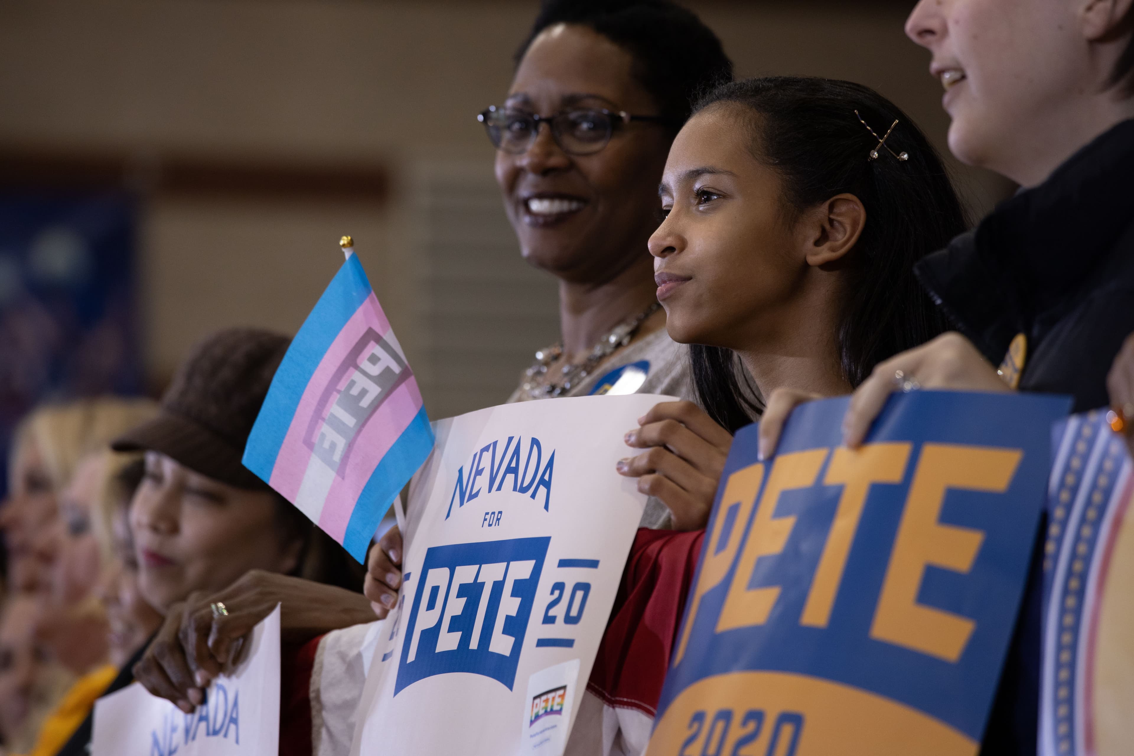 A diverse group of supporters is shown at a political rally. One person holds a sign reading "Nevada for Pete 2020," while another holds a "Pete 20" campaign sign. A person in the background holds a flag with pastel blue, pink, and white stripes.