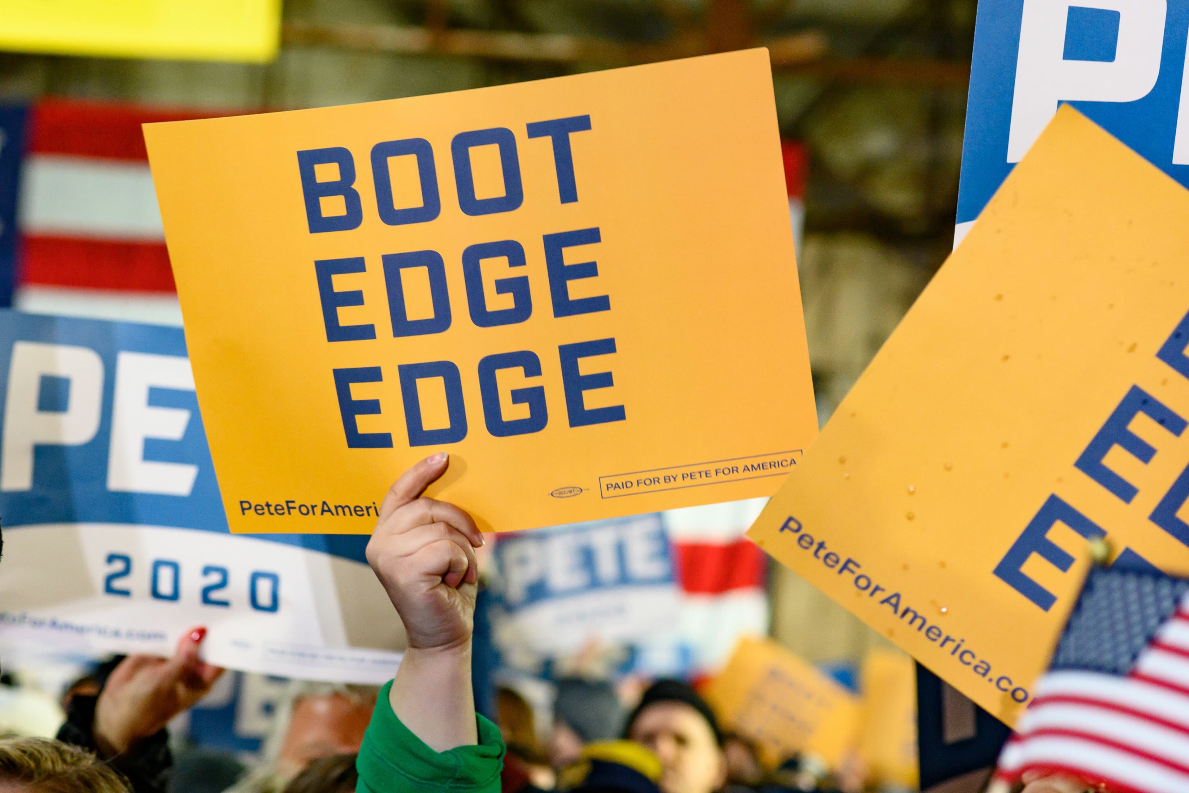 A crowd holds up a yellow sign reading "BOOT EDGE EDGE" at a political rally. Other signs are partially visible, including one with "2020" and American flag patterns. The atmosphere is lively and energetic.