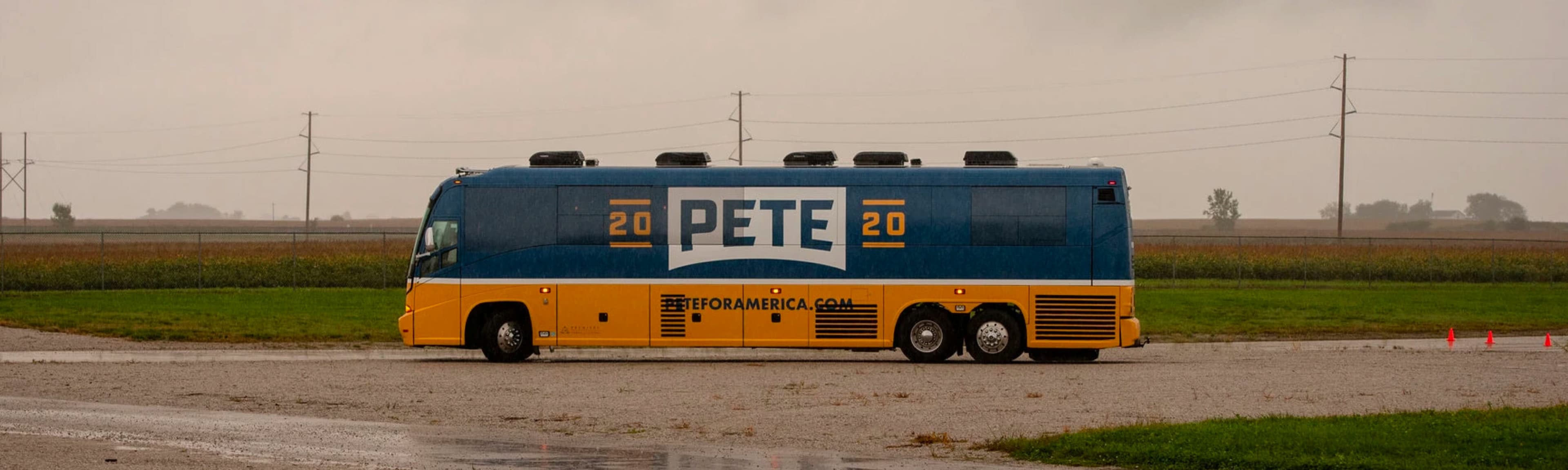 A large campaign bus with "20 Pete 20" prominently displayed on the side is parked in a rural area. The bus is blue and yellow and has a website address "TROMAMERICA.COM" written on the side. The landscape features open fields and power lines against a cloudy sky.