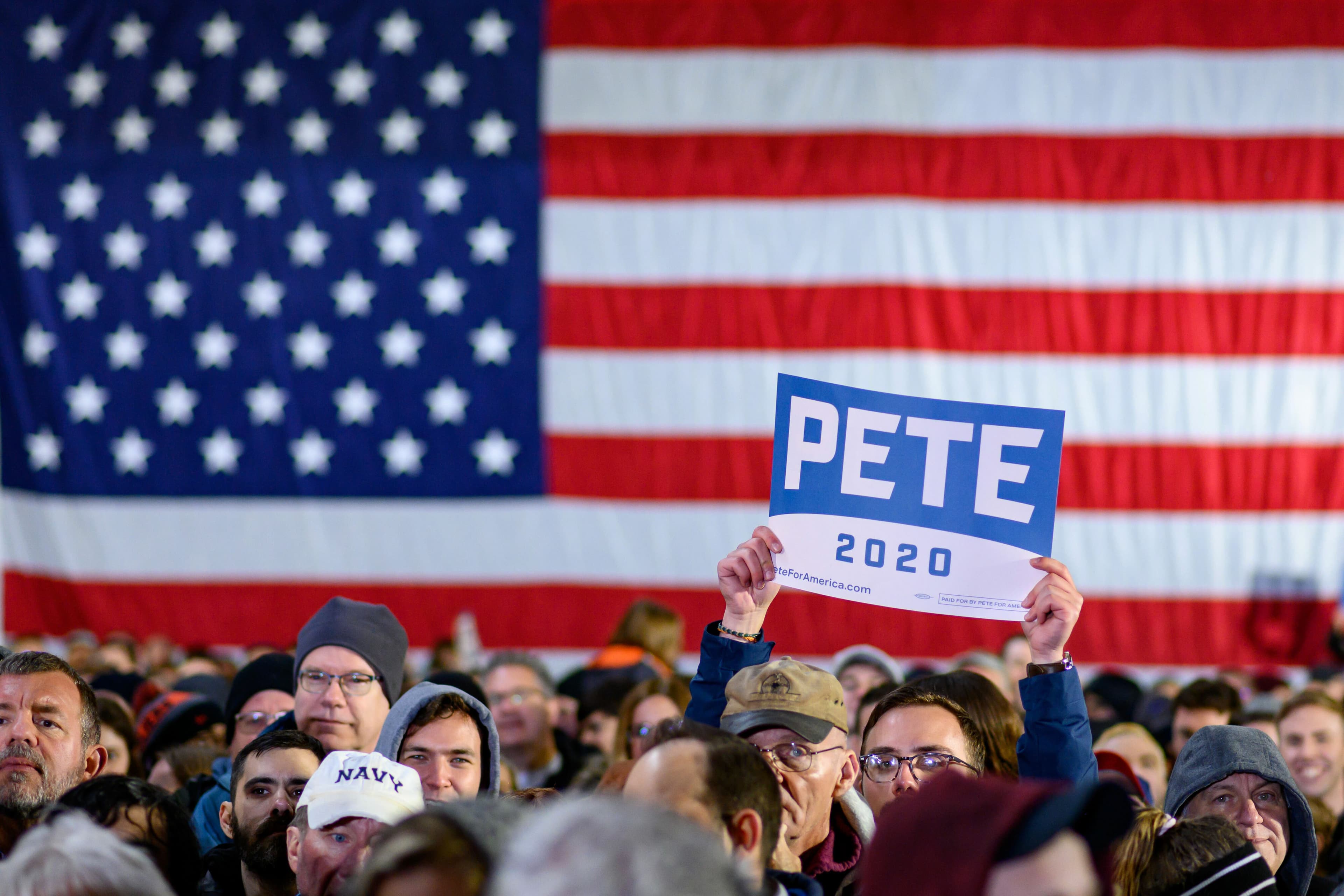 A crowd of people, dressed in winter clothing, stands in front of a large American flag. One person in the middle holds up a sign that reads "PETE 2020" above the crowd.