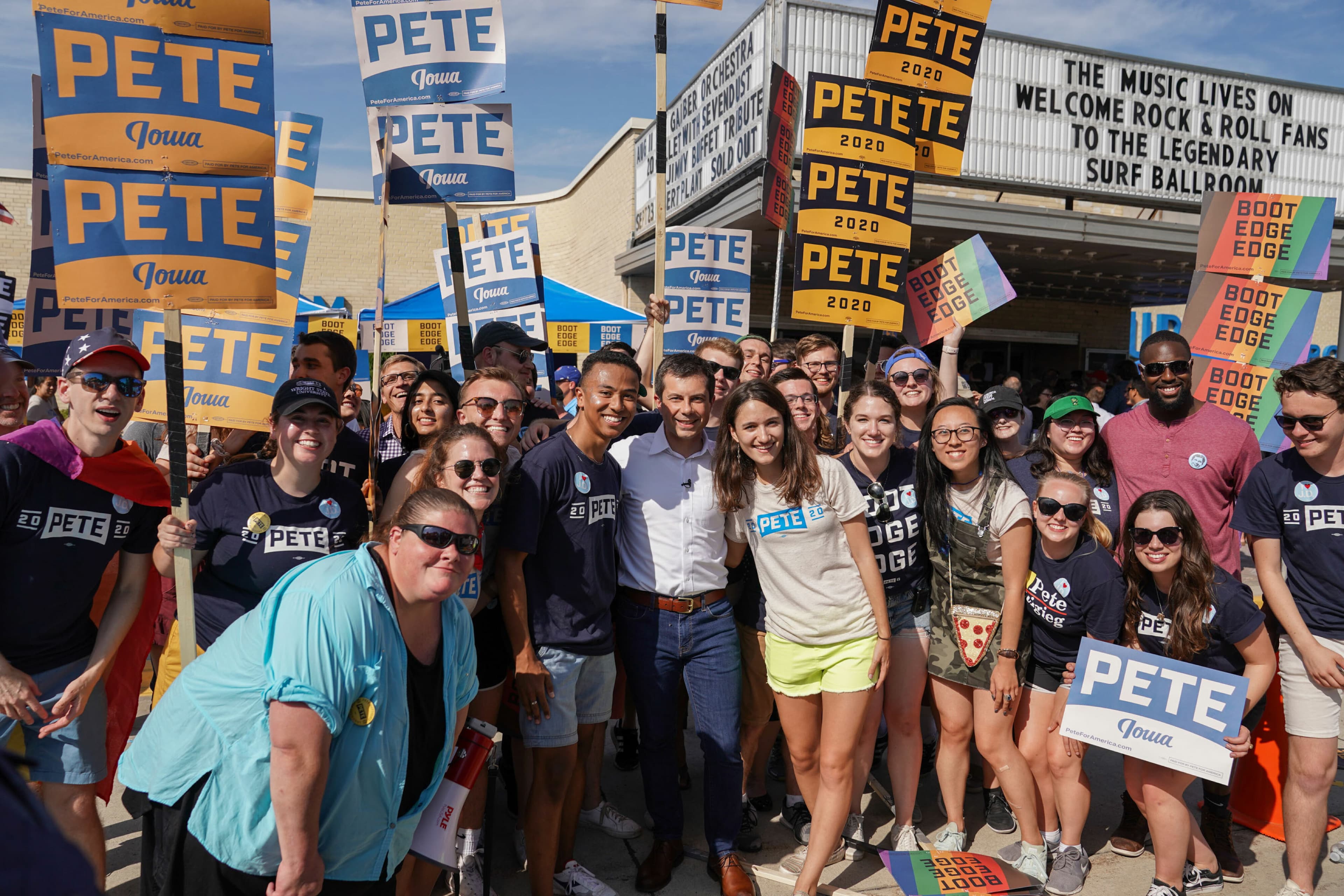 A large group of people stands outside a building holding "Pete 2020" and "Boot Edge Edge" signs. They are smiling and posing for a photo, wearing shirts and buttons that also support Pete. The marquee behind them reads, "The music lives on, Welcome Rock & Roll Fans To The Legendary Surf Ballroom.