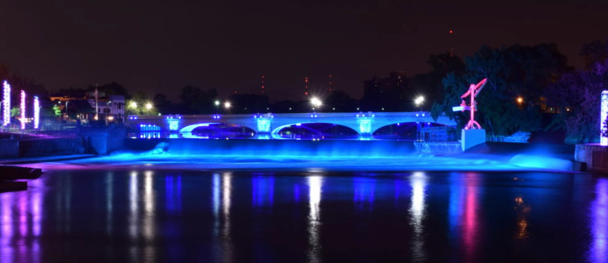 A night view of a bridge illuminated with vibrant blue and purple lights, reflecting off the calm water below. Trees and buildings can be seen in the background, and additional light sources create a colorful and serene ambiance.