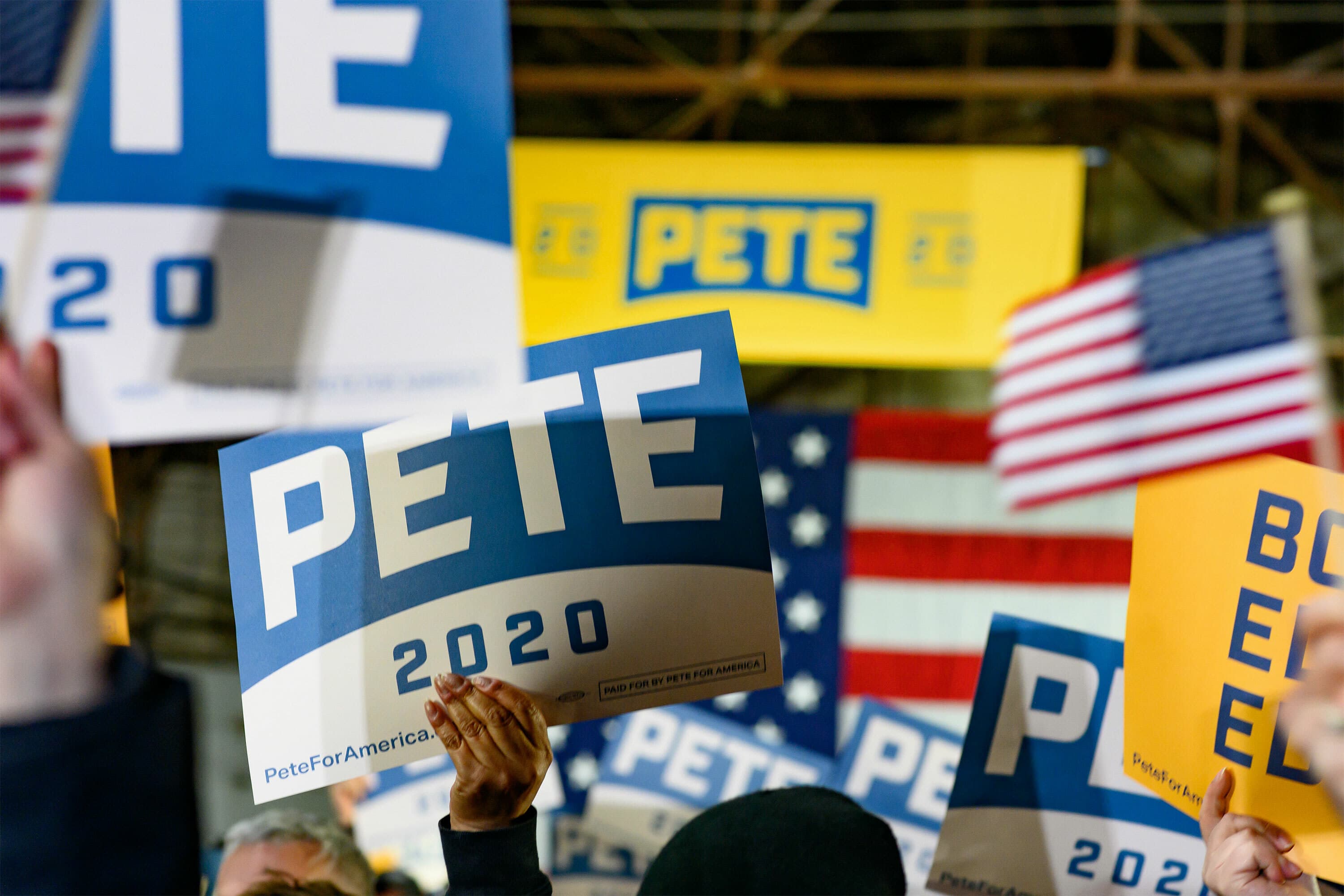 A crowd of people holding up signs reading "PETE 2020" at a rally. The background features American flags and a large banner with "PETE" and "2020" visible. The atmosphere suggests a political campaign event.